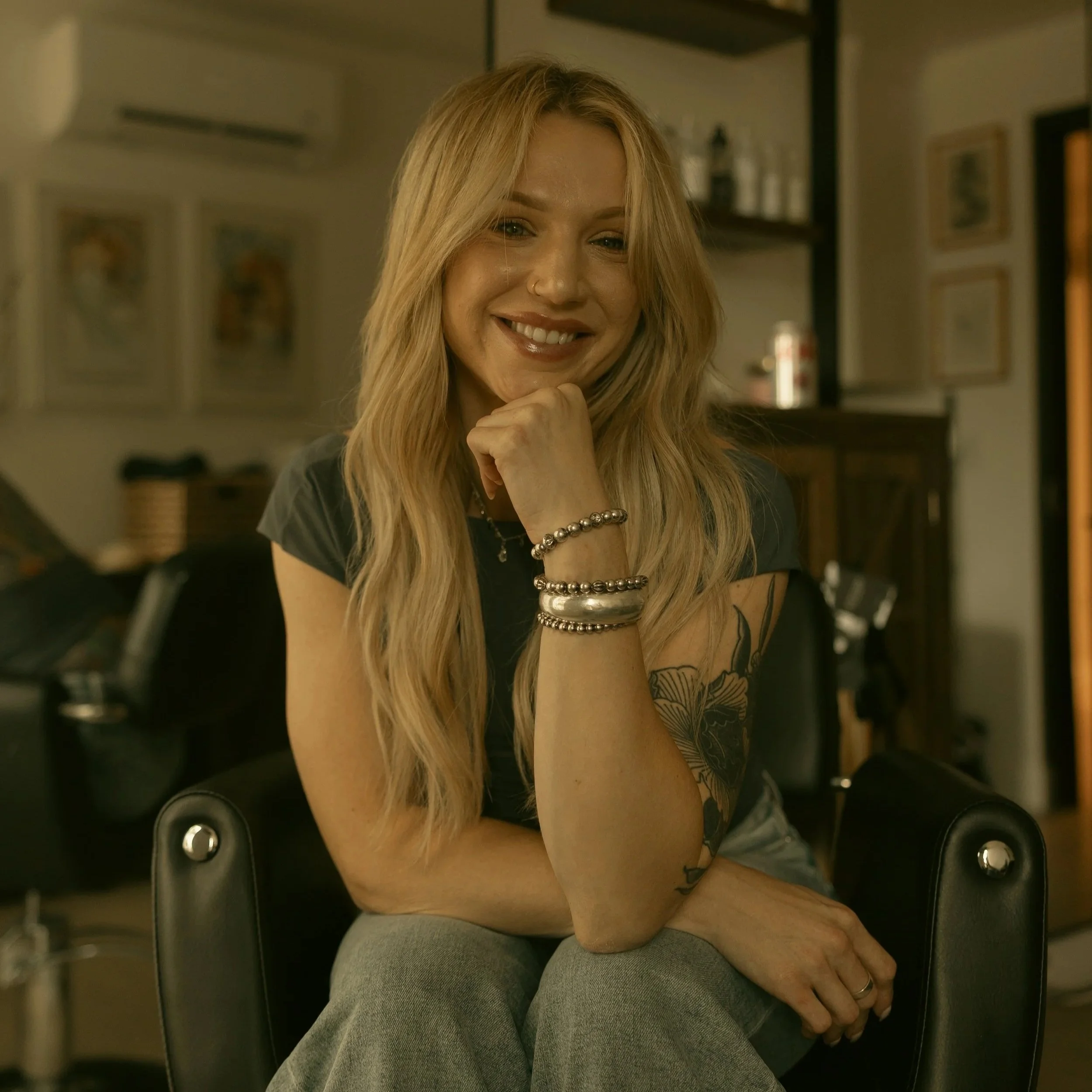 The owner of Solstice Hair Studio, Christian Taylor, with long blonde hair, wearing a nose ring, jewelry on her wrist and a tattoo on her arm, sitting on the floor of a room with framed pictures on the wall, in an intimate candid moment.