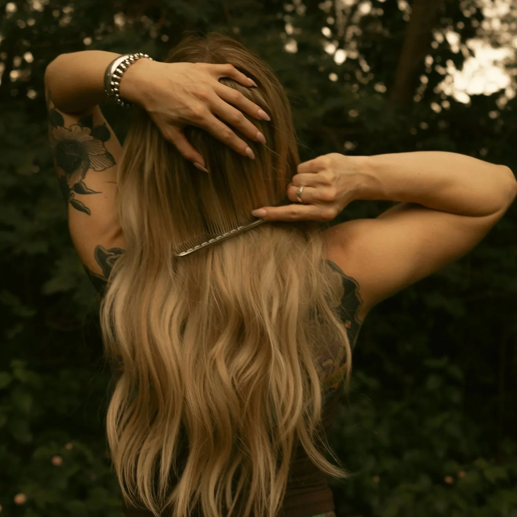 A woman with long, blonde hair applying hairstyling or grooming in an outdoor setting, with trees in the background.