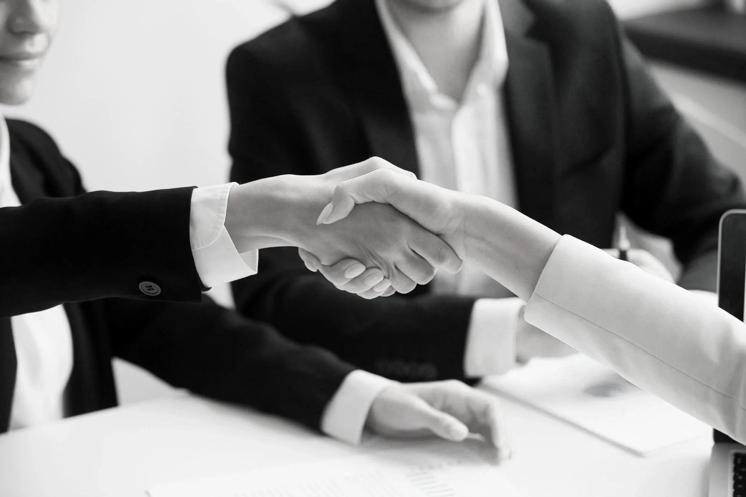 Close-up of two people in business attire shaking hands over a white table in a professional setting.