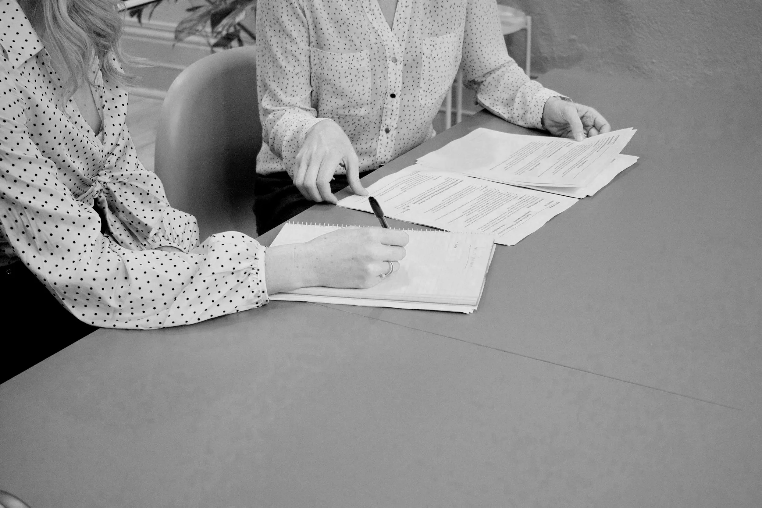 Two women sitting at a table reviewing and taking notes on several sheets of paper, with one woman holding a pen.