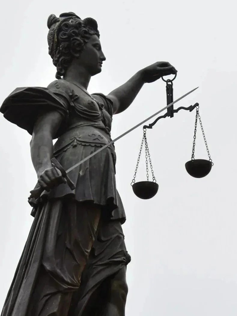 Statue of a woman with long hair styled in curls, wearing a dress and holding scales in her right hand, symbolizing justice, against an overcast sky.