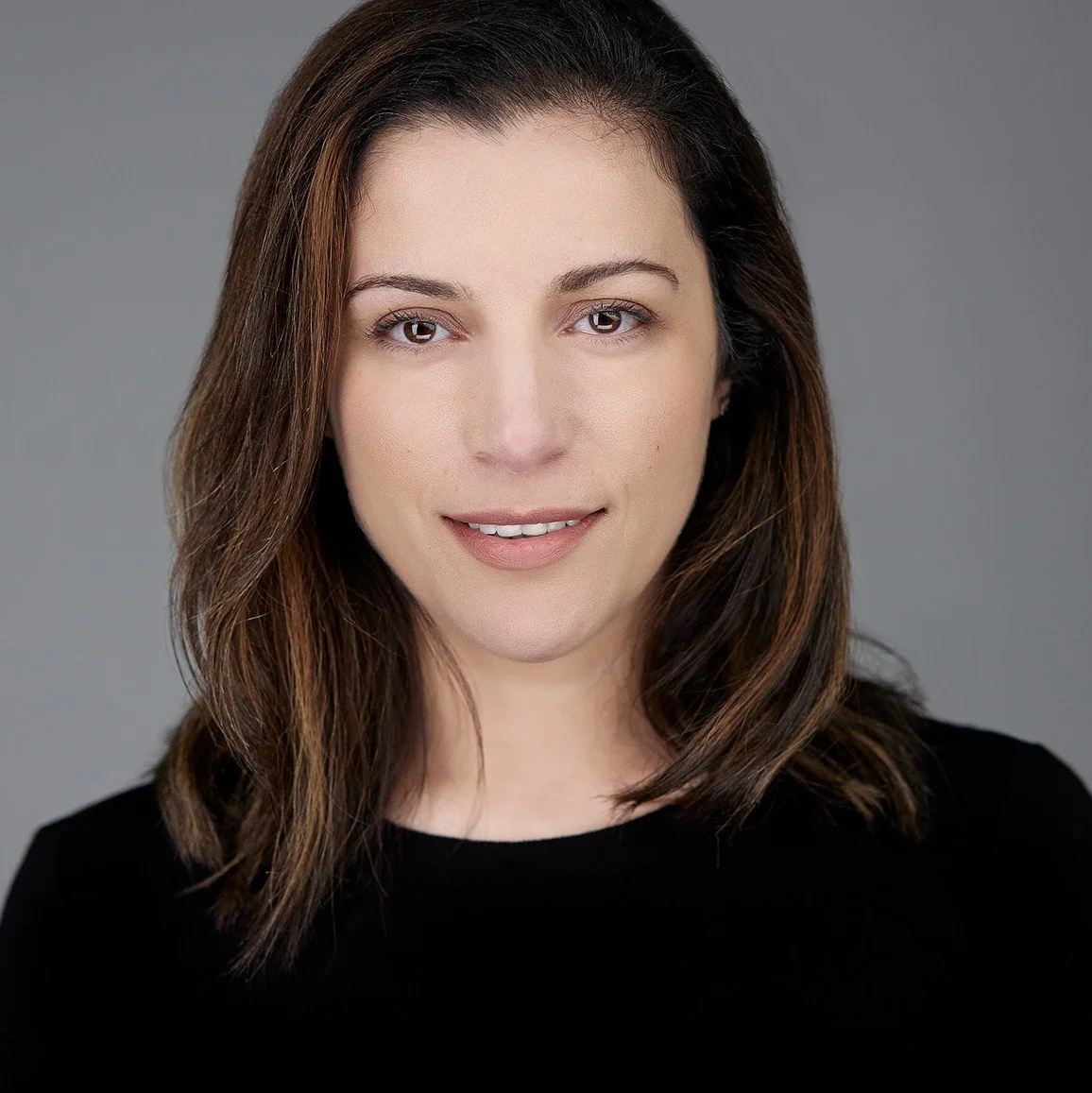 Close-up headshot of a woman with brown hair, wearing a black top, smiling softly against a neutral gray background.