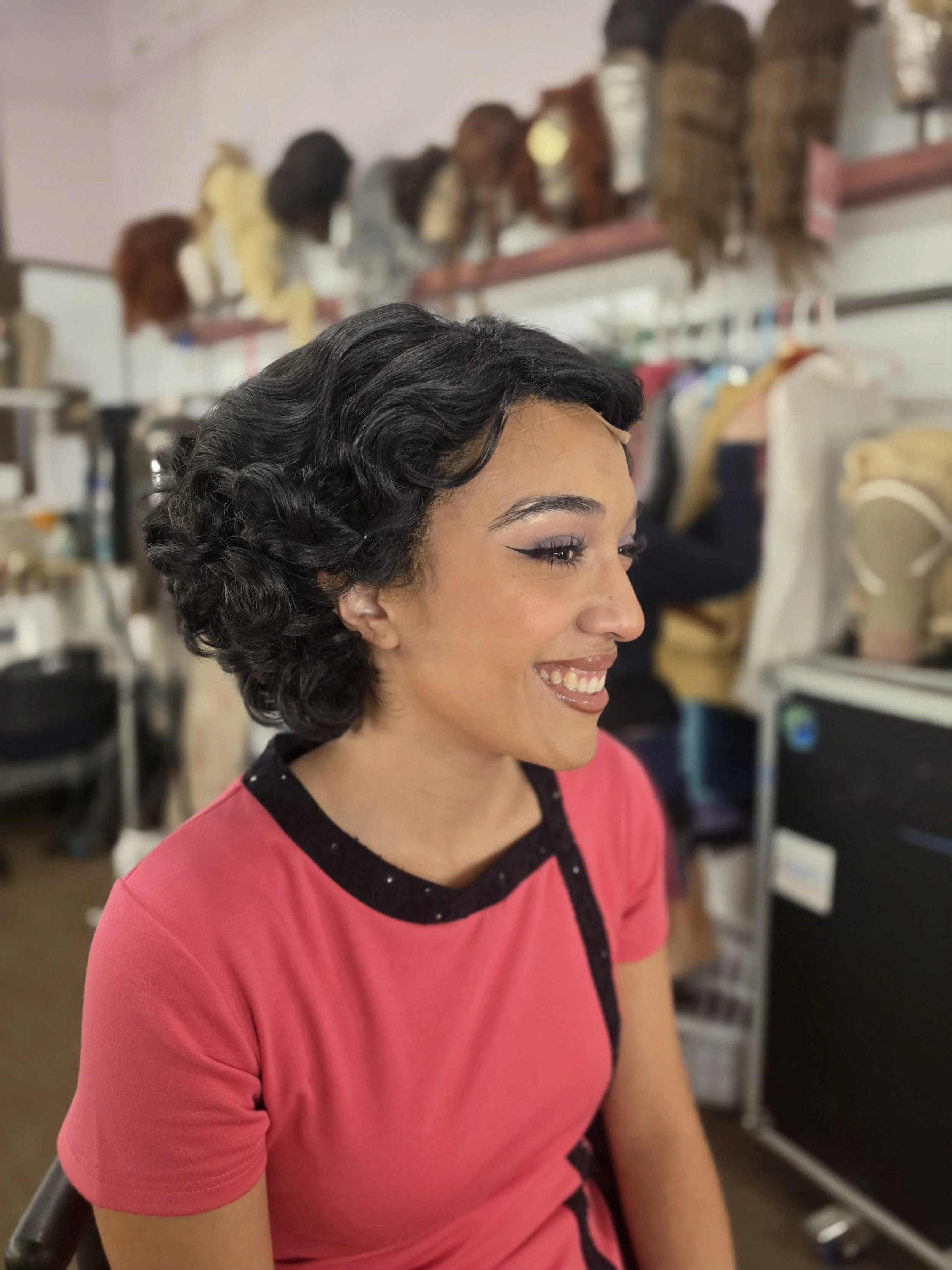 Side profile of a woman with short black curly hair smiling, wearing a pink top with black accents, in a room with wig stands and styling tools in the background.