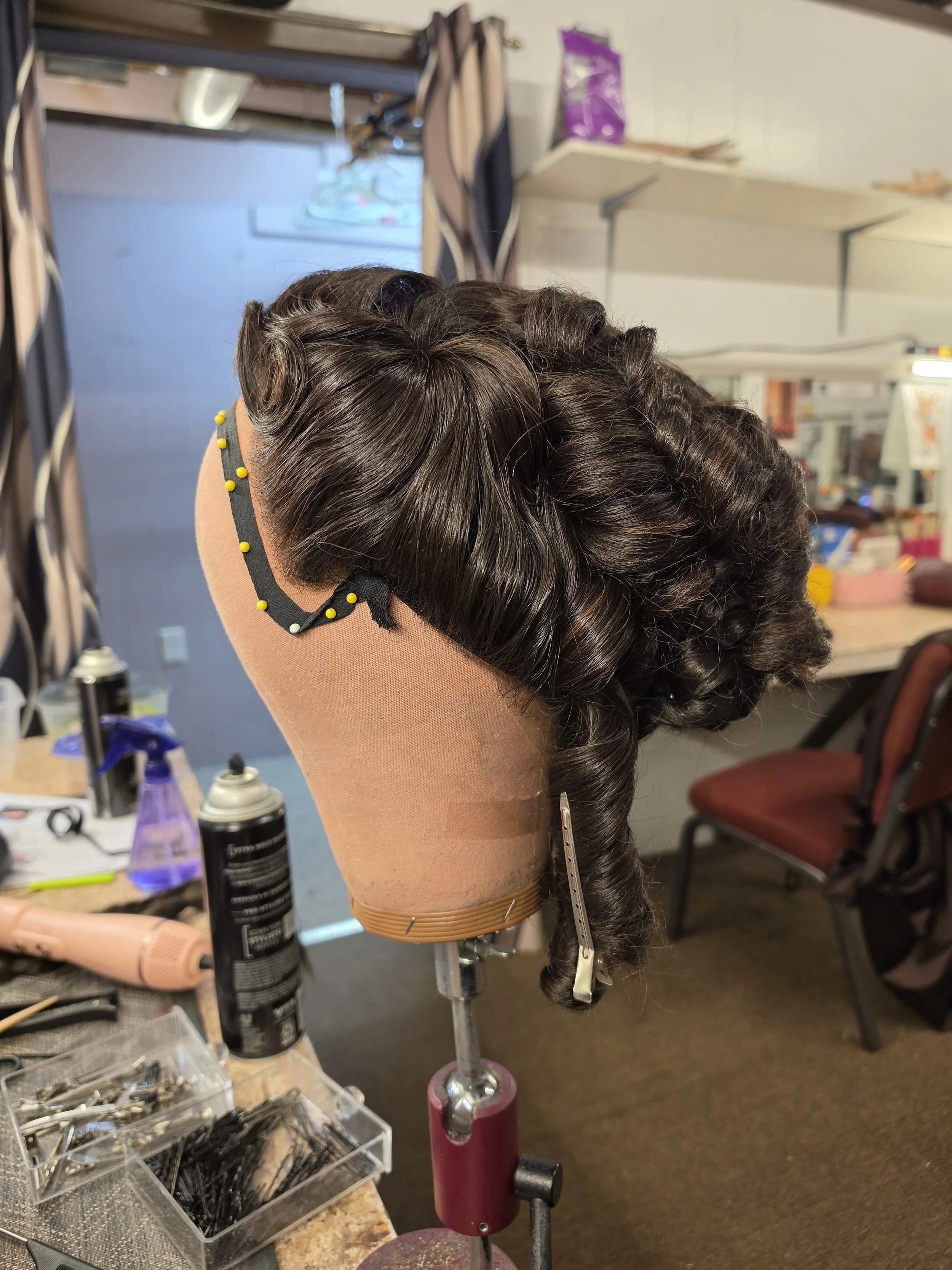 A hair mannequin head with styled dark brown hair on a salon table, surrounded by hair tools and supplies in a salon or training room.