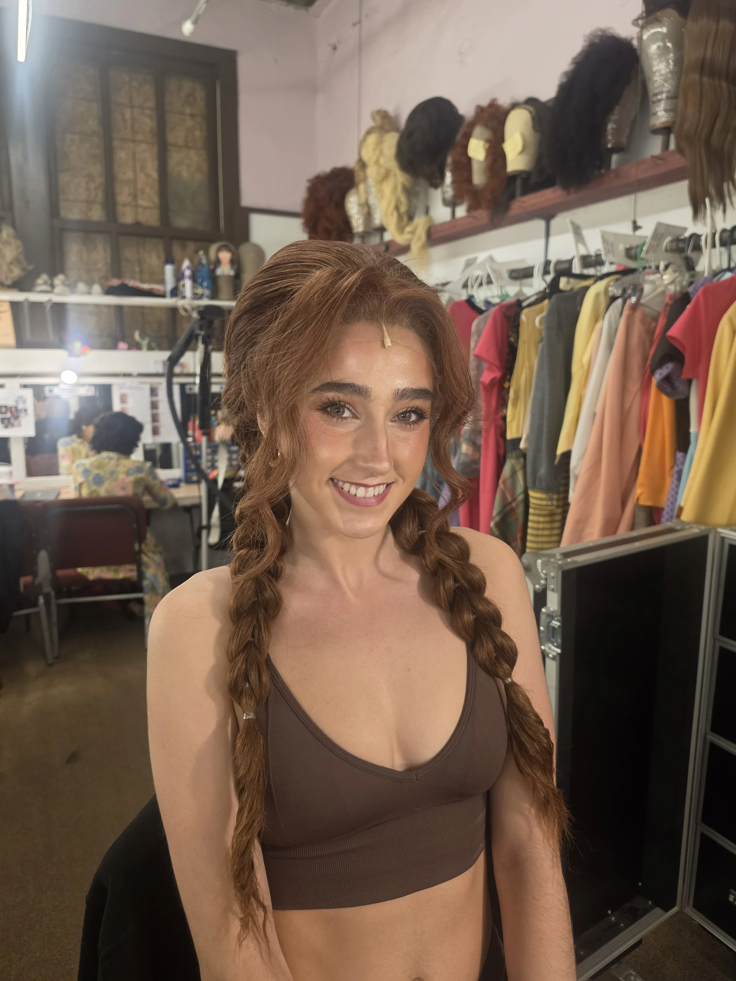 A woman with red hair styled in long, loose curls is smiling at the camera. She is wearing a brown sports bra and has a hairpin in her hair. The background appears to be a makeup or costume room with wigs on shelves, clothing hanging on racks, and va
