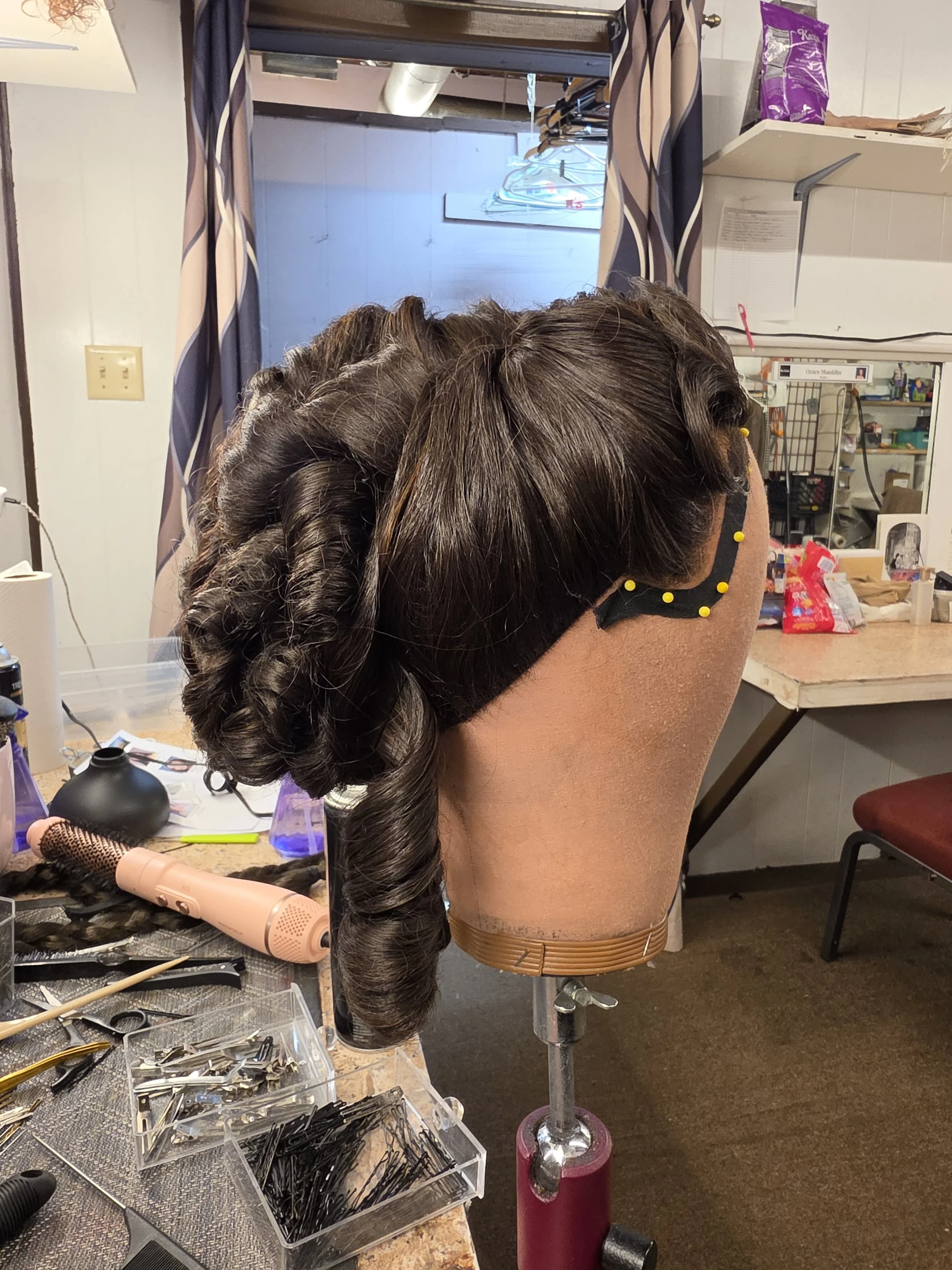 A sewing or hairstyling workspace with a mannequin head displaying styled curly hair, surrounded by various tools, hairpins, and products on a cluttered table. In the background, there are hangers, a mirror, and some storage shelves.