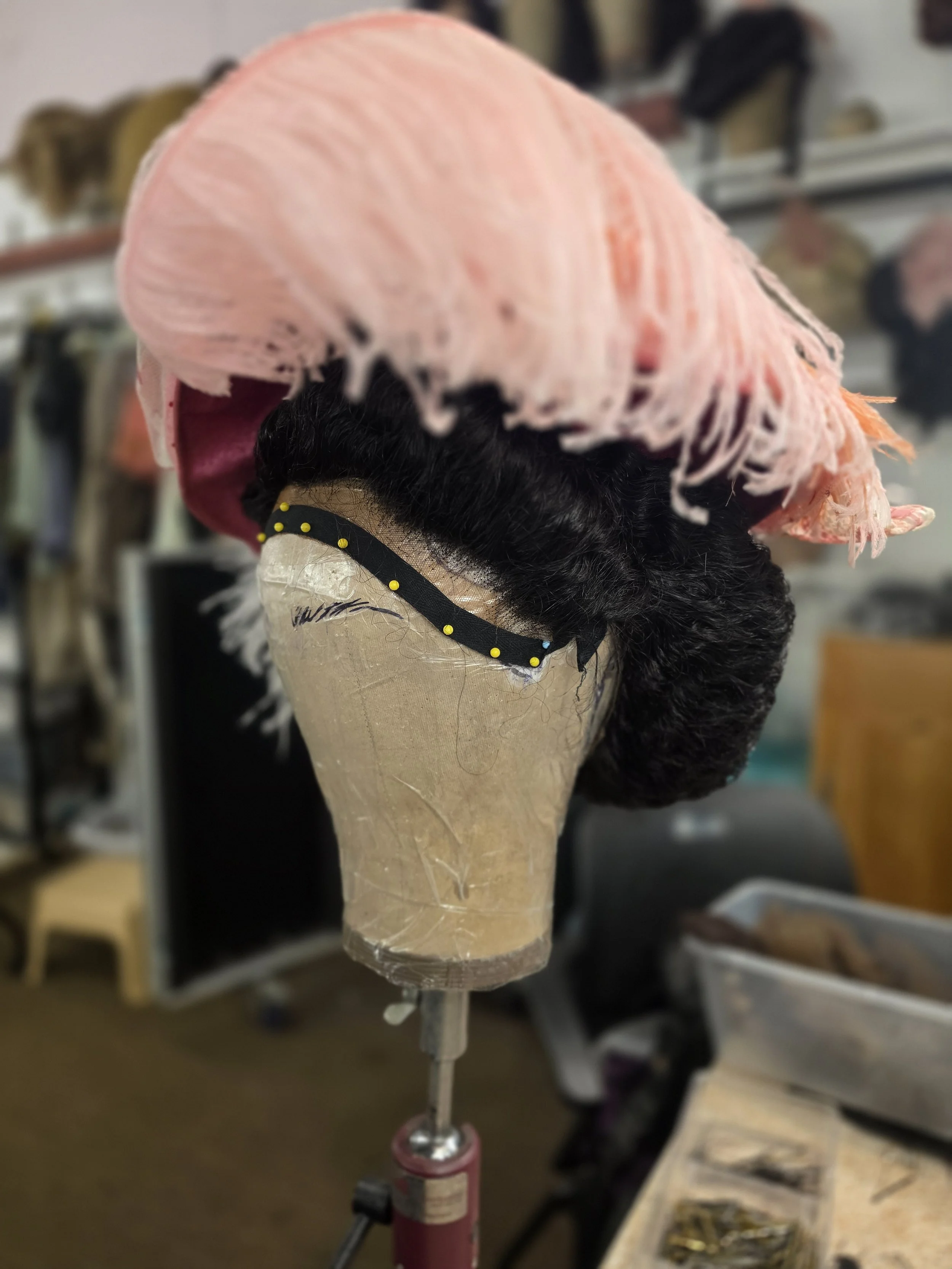 A mannequin head with black curly hair wearing a light pink frayed hat, placed on a worktable in a workshop.