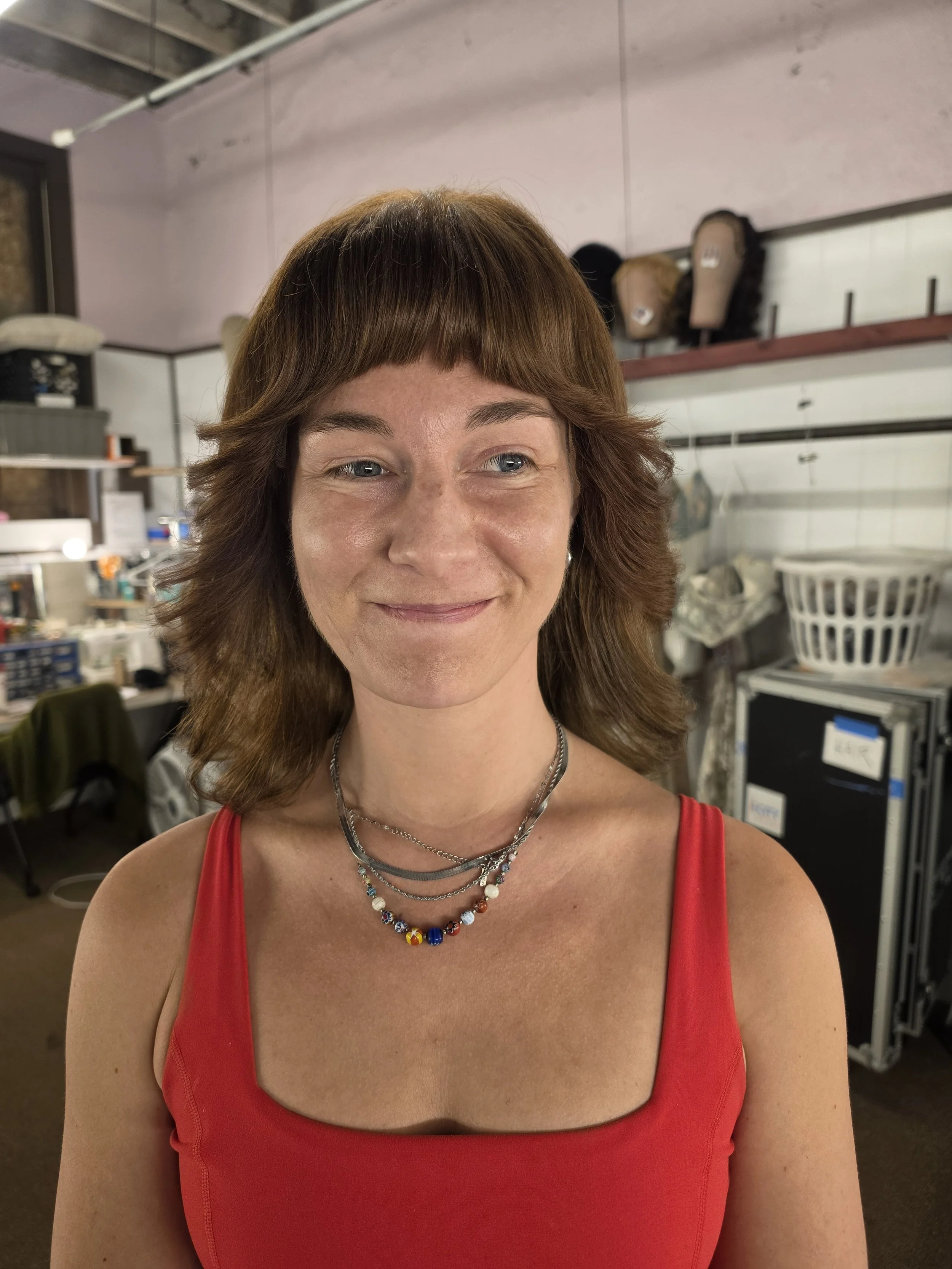 A woman with brown hair and blue eyes is smiling while taking a selfie indoors. She is wearing a red sleeveless top and colorful necklaces. The background appears to be a storage or utility room with shelves, boxes, and various items.
