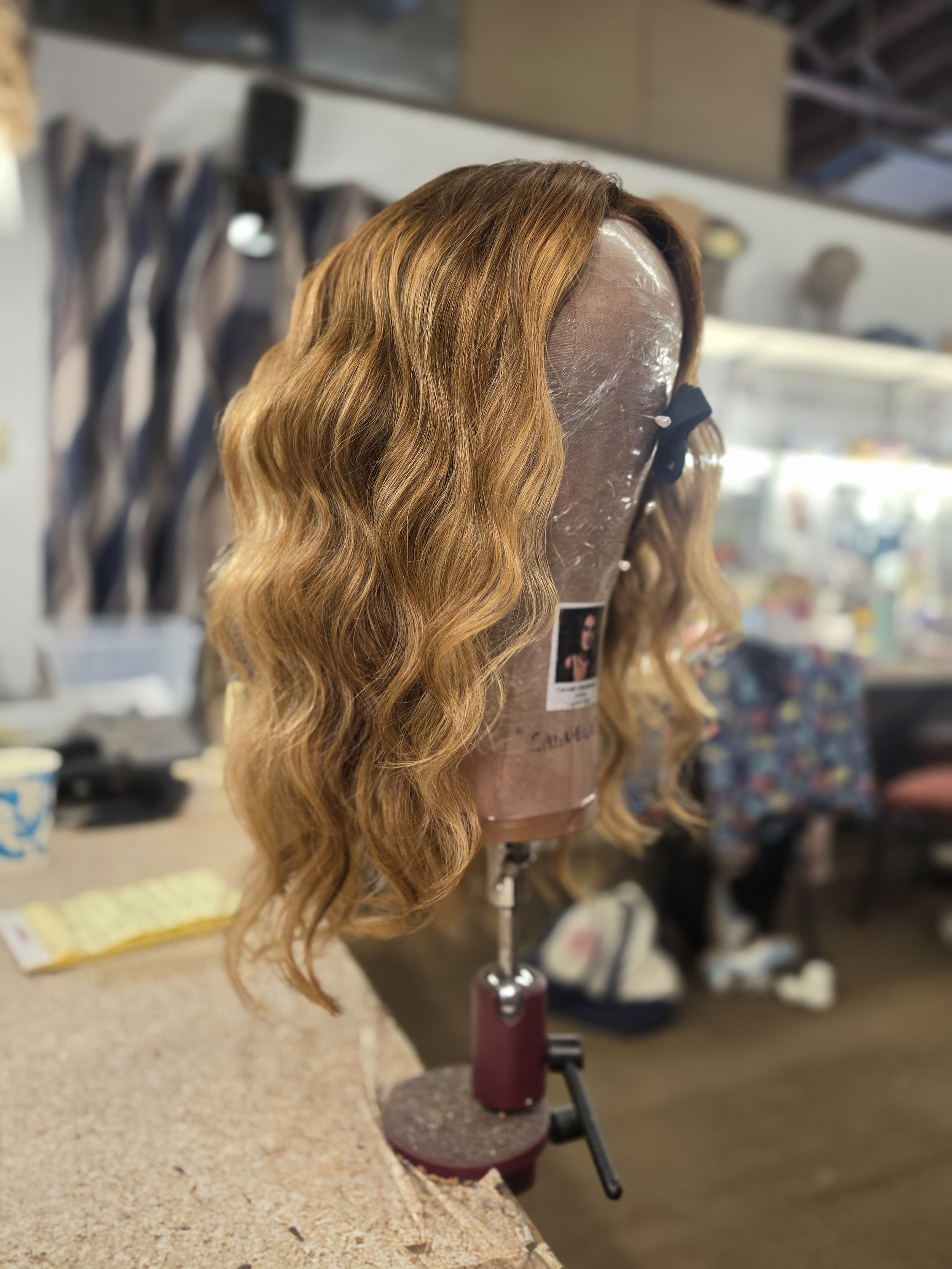 A wig with wavy, reddish-brown hair on a mannequin head, attached with a black hair clip, displayed on a stand in a store or salon environment.