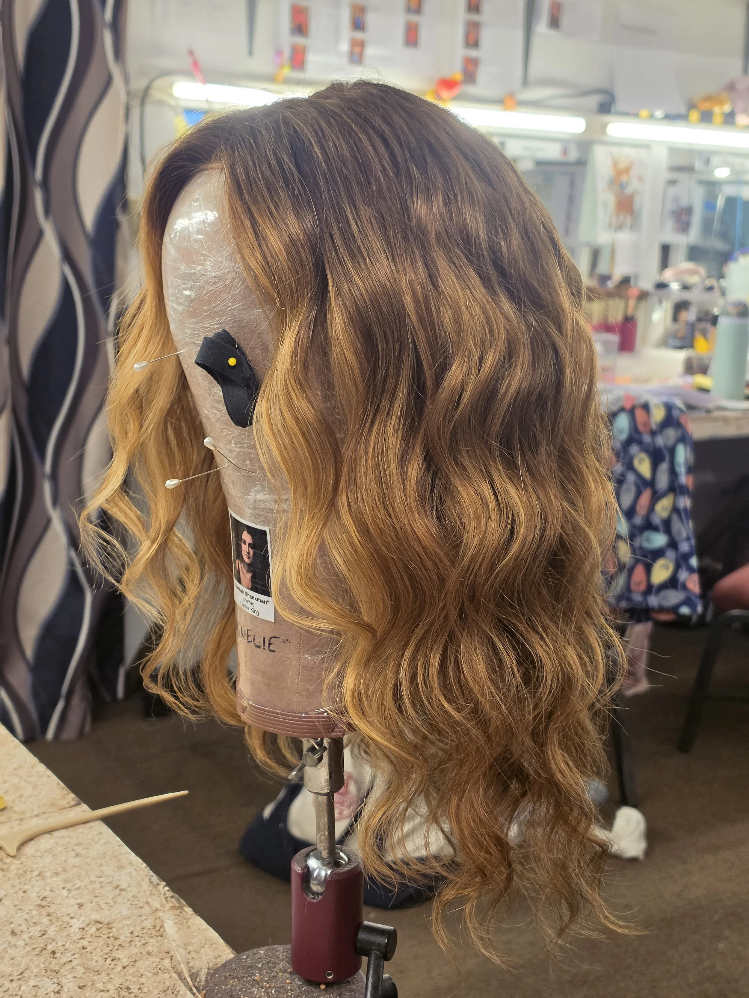 A wig styled with wavy, two-toned hair, light brown at the roots fading into darker brown, displayed on a mannequin head attached to a wig stand in a room with a mirror, makeup, and hairdressing supplies in the background.