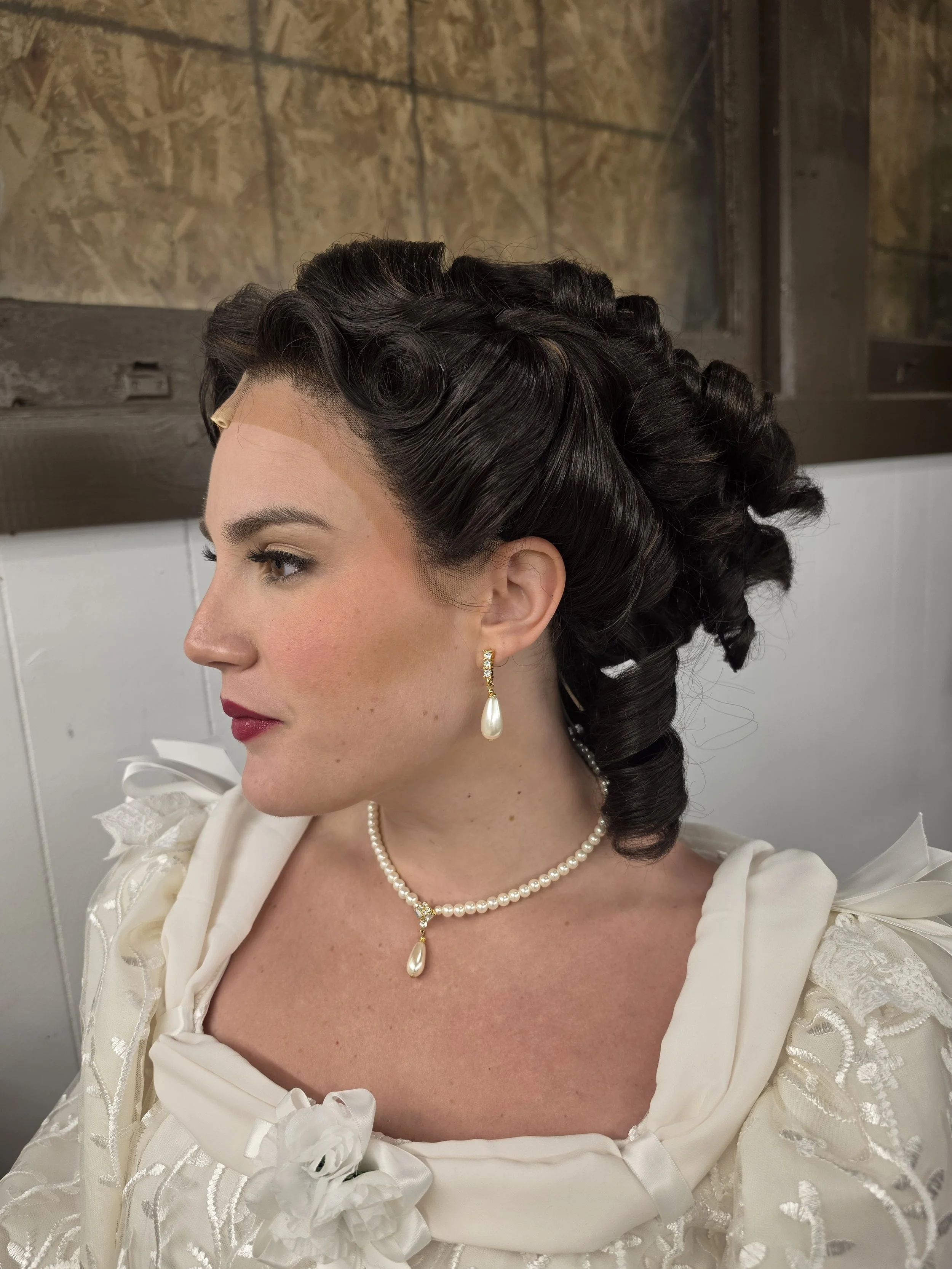 A woman dressed in vintage bridal attire, wearing a white dress with lace and floral details, pearl necklace and earrings, styled dark hair, and natural makeup, is captured in profile against a neutral background.
