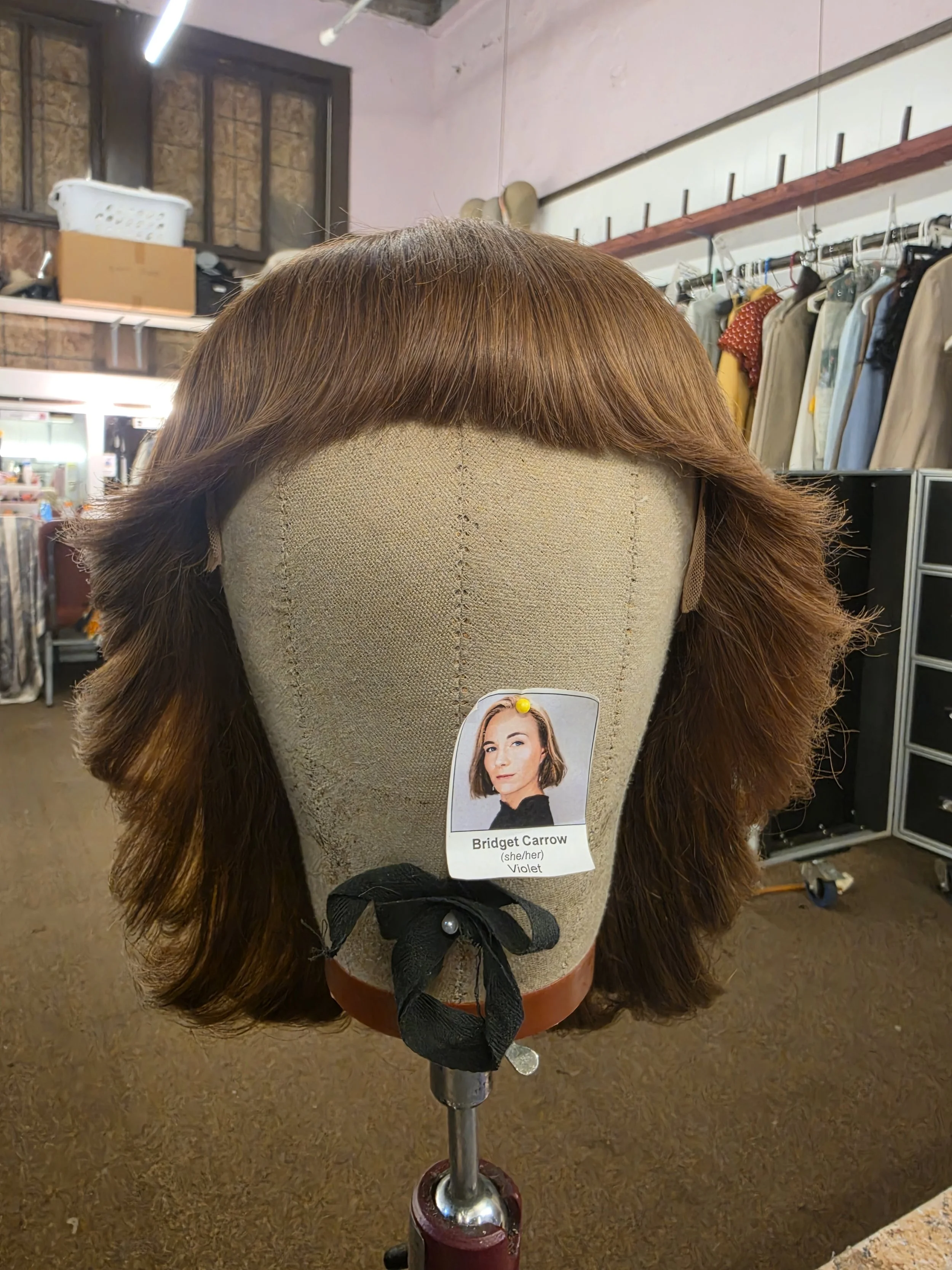 A mannequin head with a brown wig with straight bangs, labeled as Bridget Carrow, violet, and she/her pronouns, in a clothing or costume store.