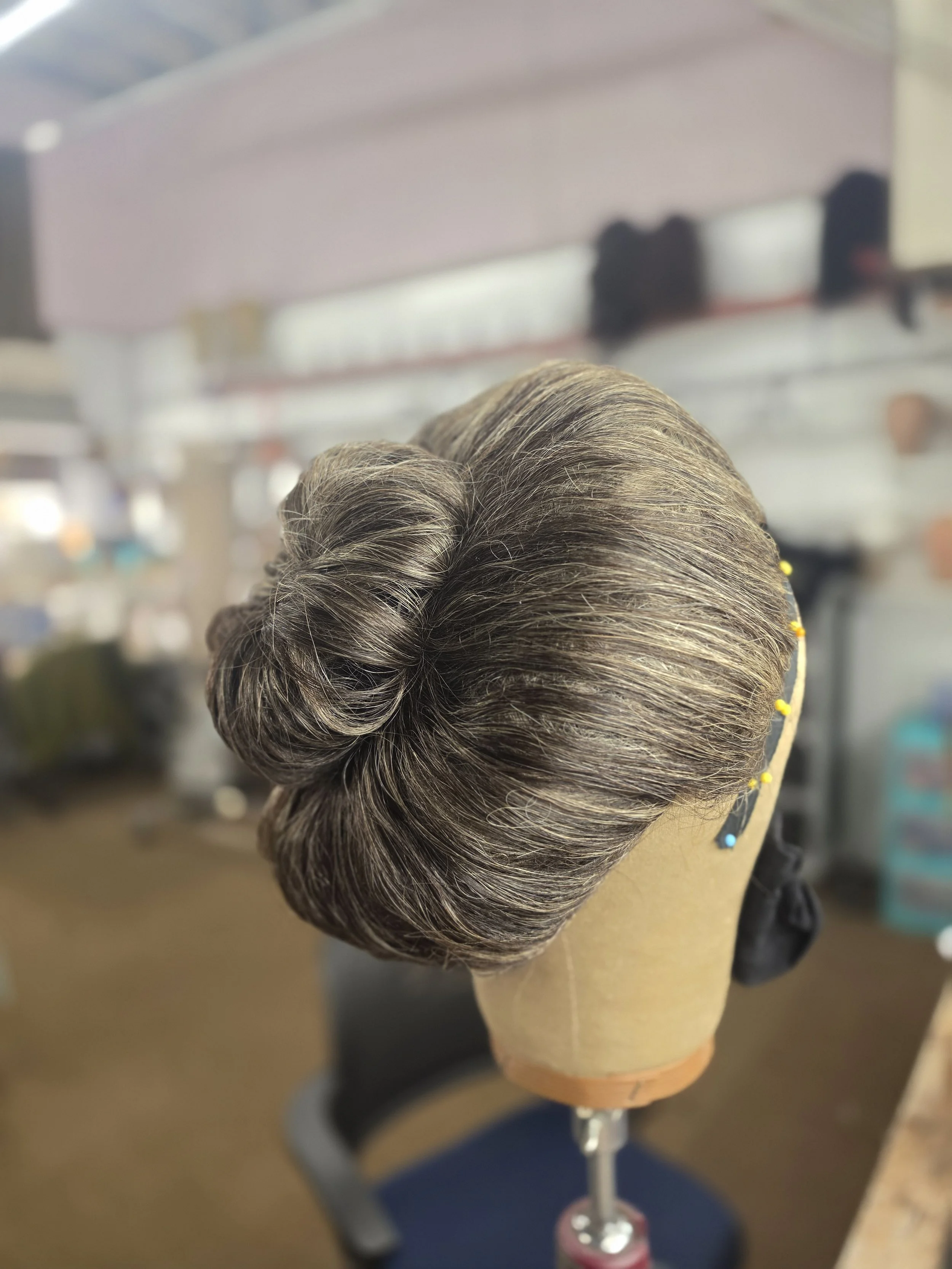 A mannequin head on a stand with a vintage-style gray hairdo, styled in a rolled-up updo with highlights, placed in a salon or studio setting.