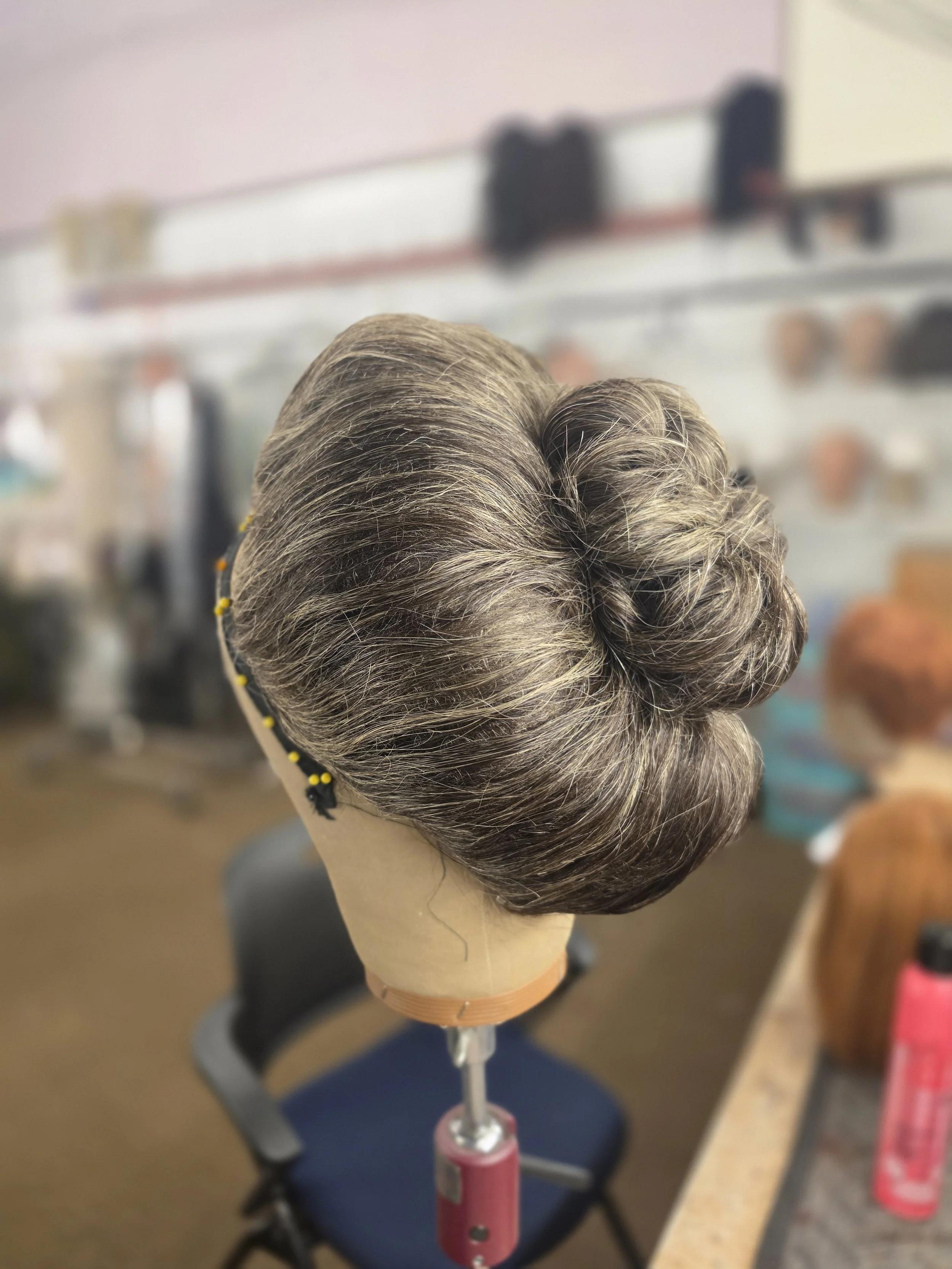 A mannequin head with gray and white styled hair, wearing a black ribbon with yellow beads, placed on a pink stand in a salon or barber shop.