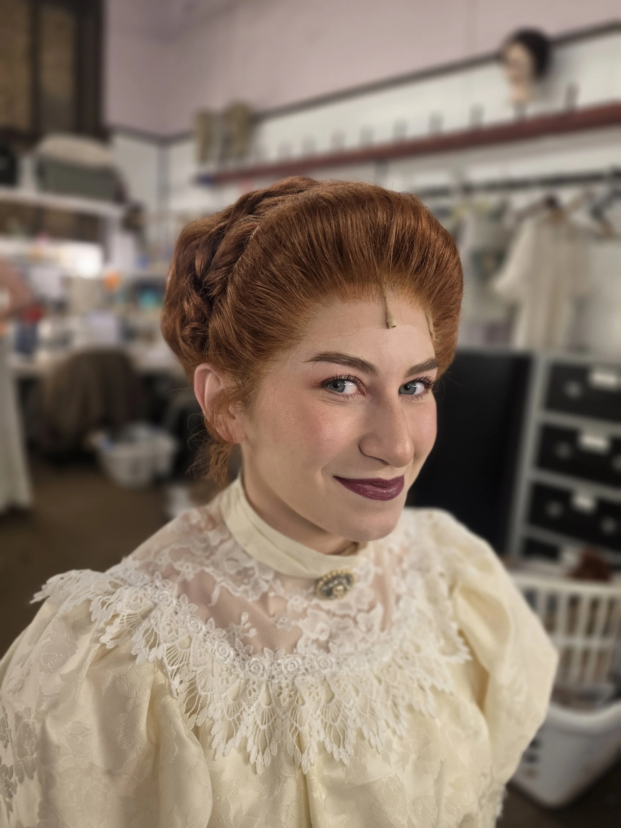 Woman with styled red hair wearing an elegant cream-colored lace dress in a room with shelves and electronic equipment.