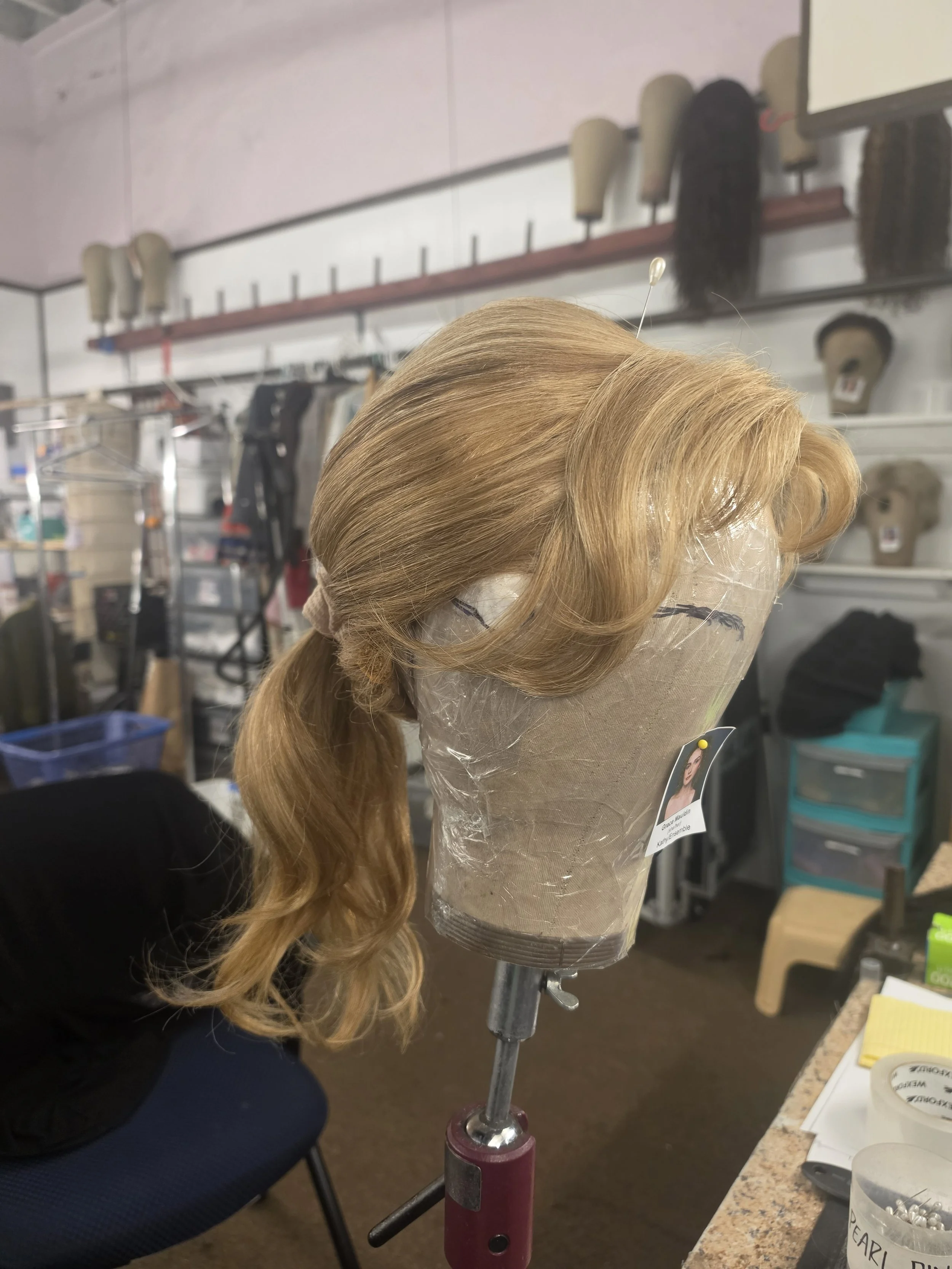 A blonde wig on a mannequin head secured with a pin, placed on a stand in a costume or wig store. Shelves with wig caps and various hairpieces are visible in the background.