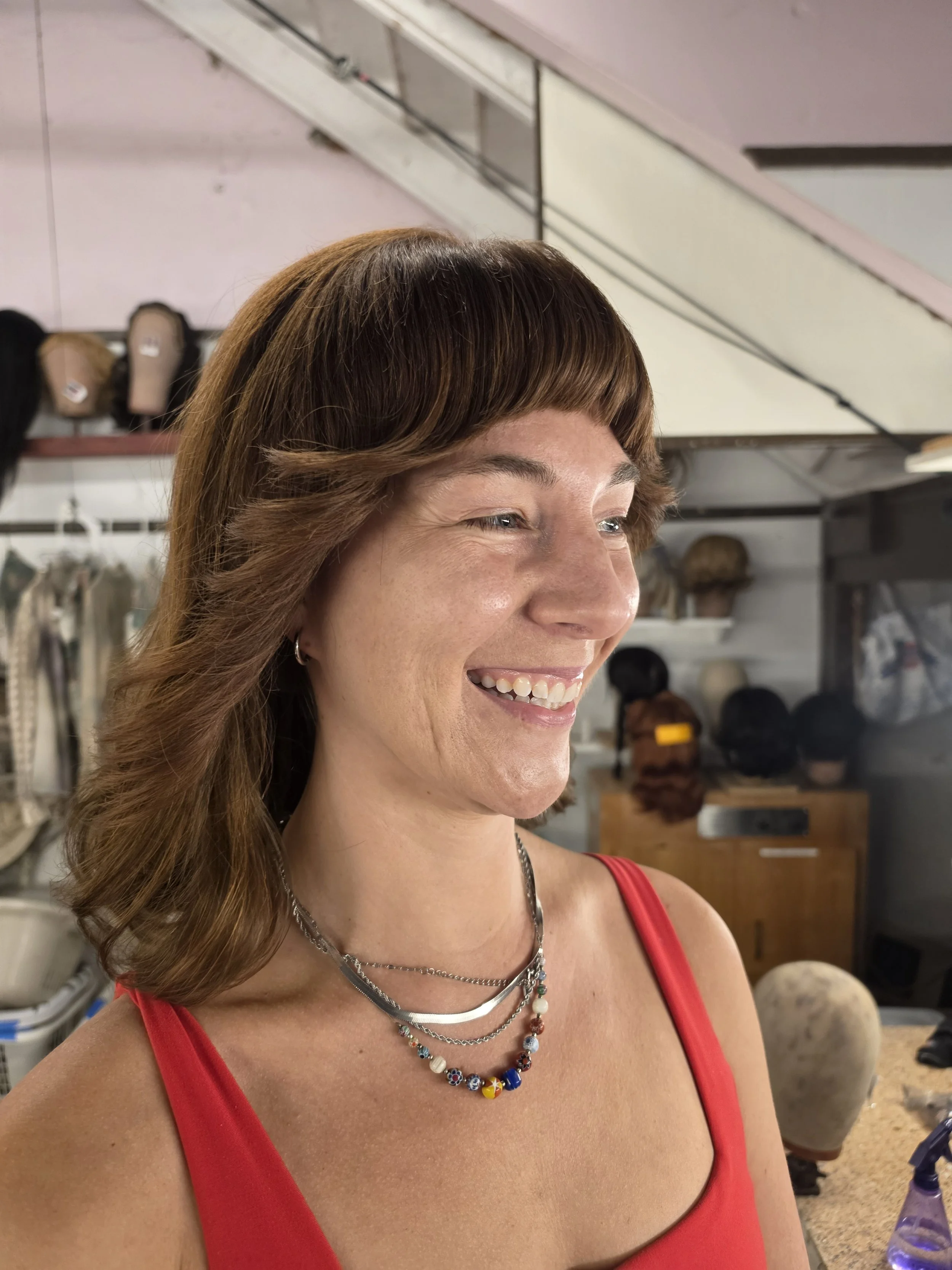 A woman with medium-length, wavy brown hair with bangs, smiling and wearing a red top, multiple layered necklaces with colorful beads, and earrings, in a setting with hats and wigs on shelves in the background.