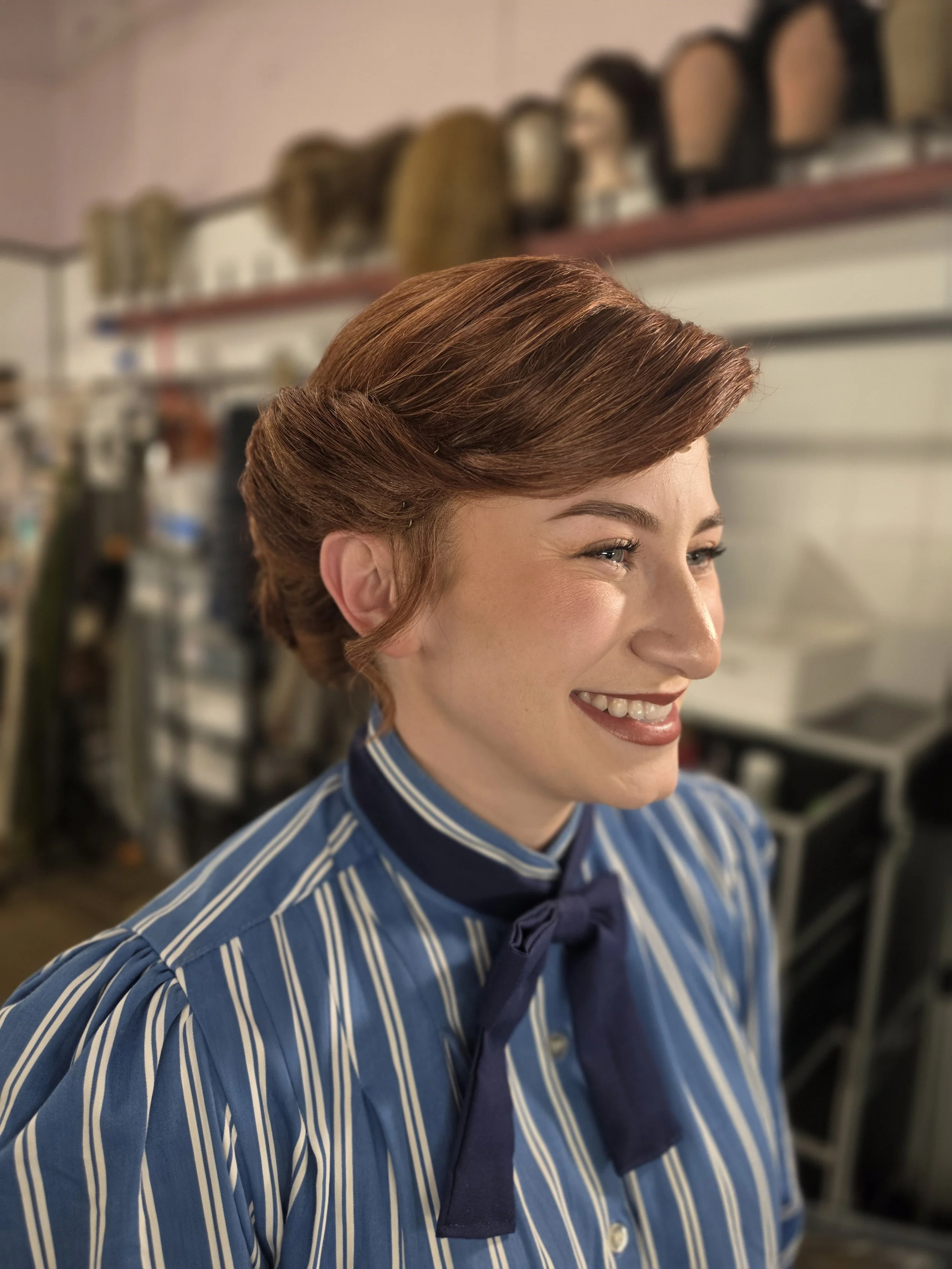 A woman with neatly styled, reddish-brown hair smiling while wearing a blue and white striped uniform with a scarf tied around her neck, in a salon or beauty school setting.