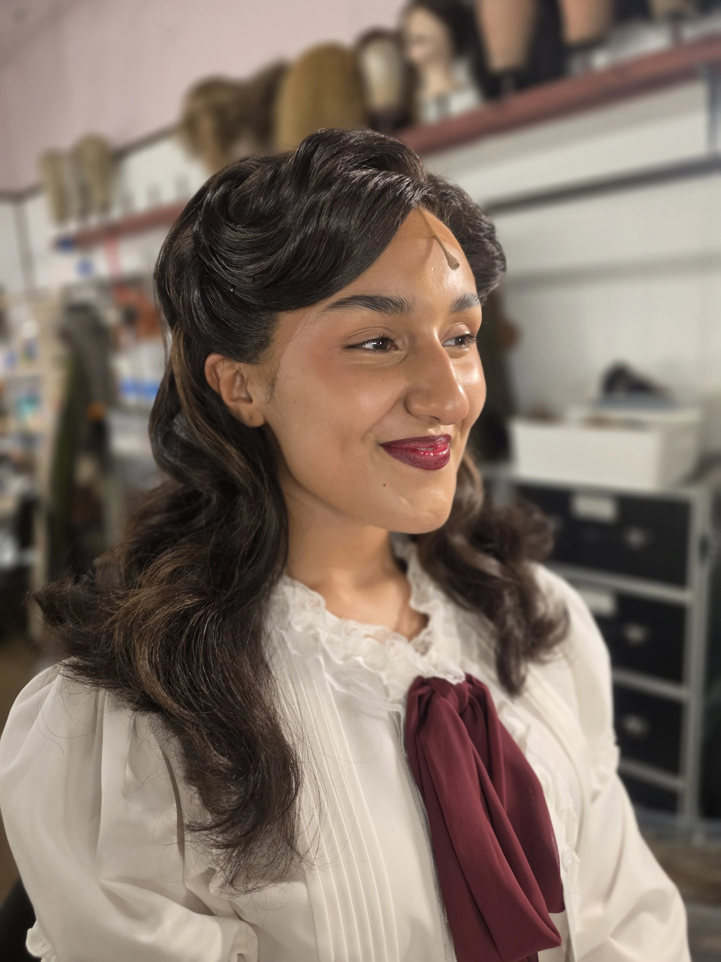 A woman with wavy dark hair styled in loose waves, wearing a white blouse with lace details and a burgundy ribbon tied at her neck, smiling gently in what appears to be a salon or dressing room with shelves and wig stands in the background.