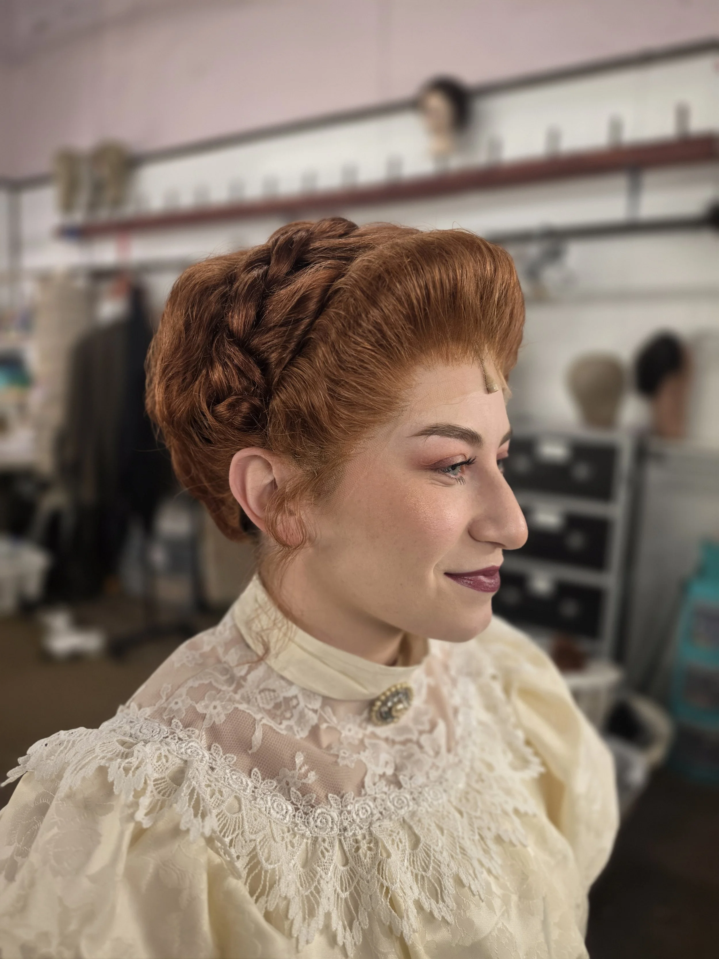 A woman with styled red hair and a vintage cream lace dress sitting in a chair, in a room with hairstyling tools and wig heads in the background.