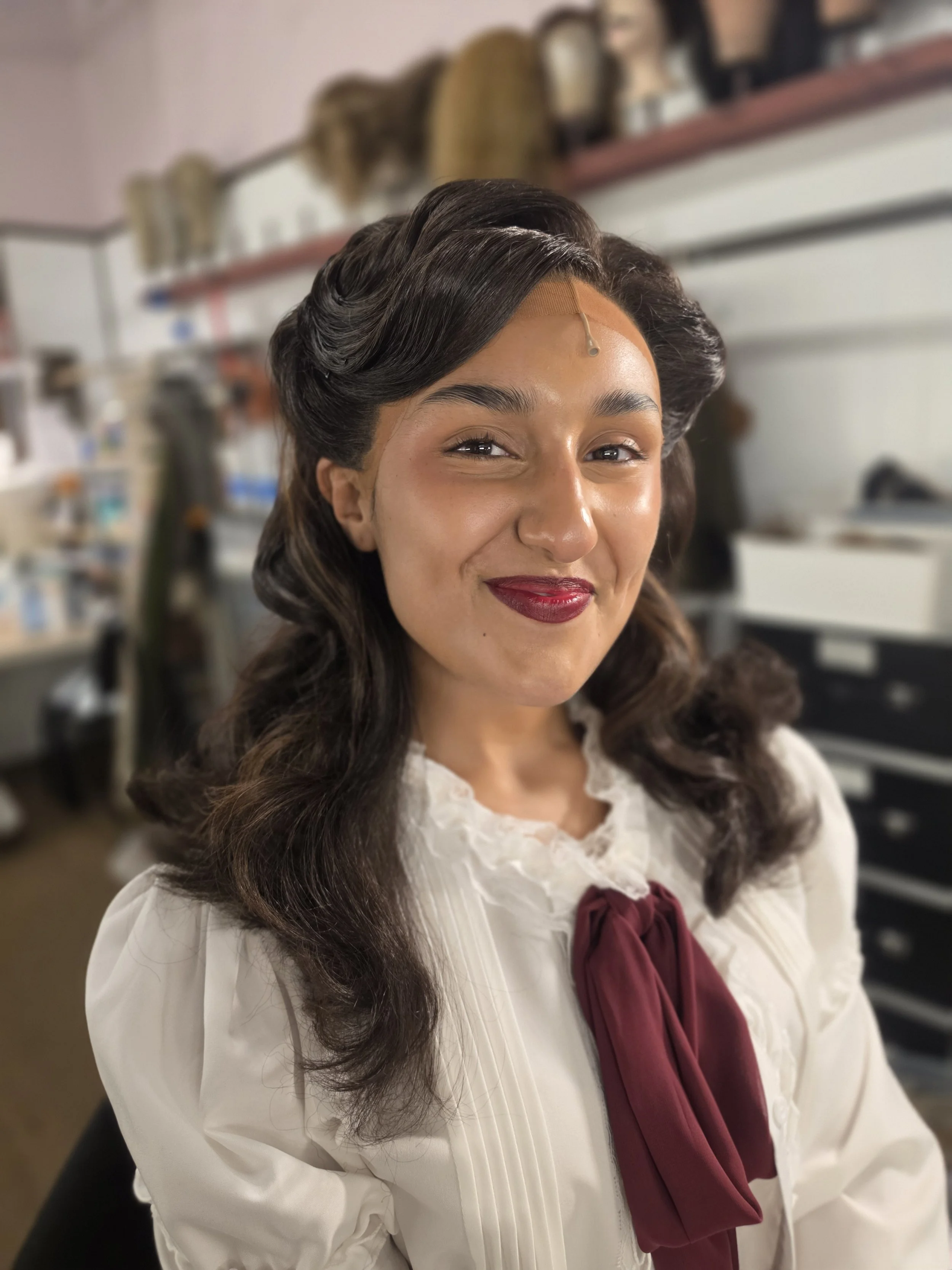 A woman with styled, wavy dark hair, wearing a white blouse with lace details and a burgundy bow, smiling in a room with shelves and various items in the background.
