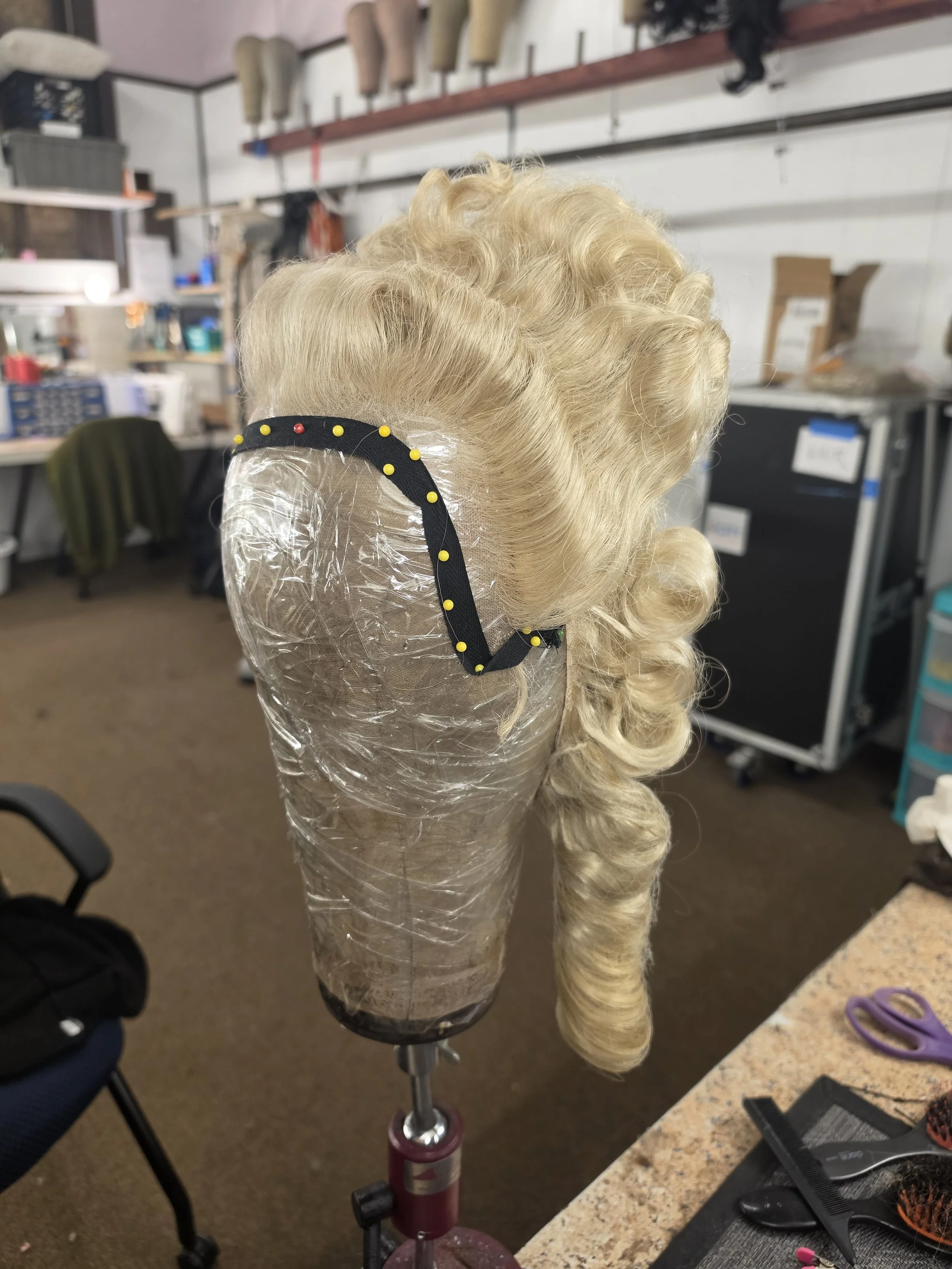 A mannequin head with long, curly blonde hair secured on a salon wig stand, set in a salon or workshop environment with shelves, tools, and supplies in the background.
