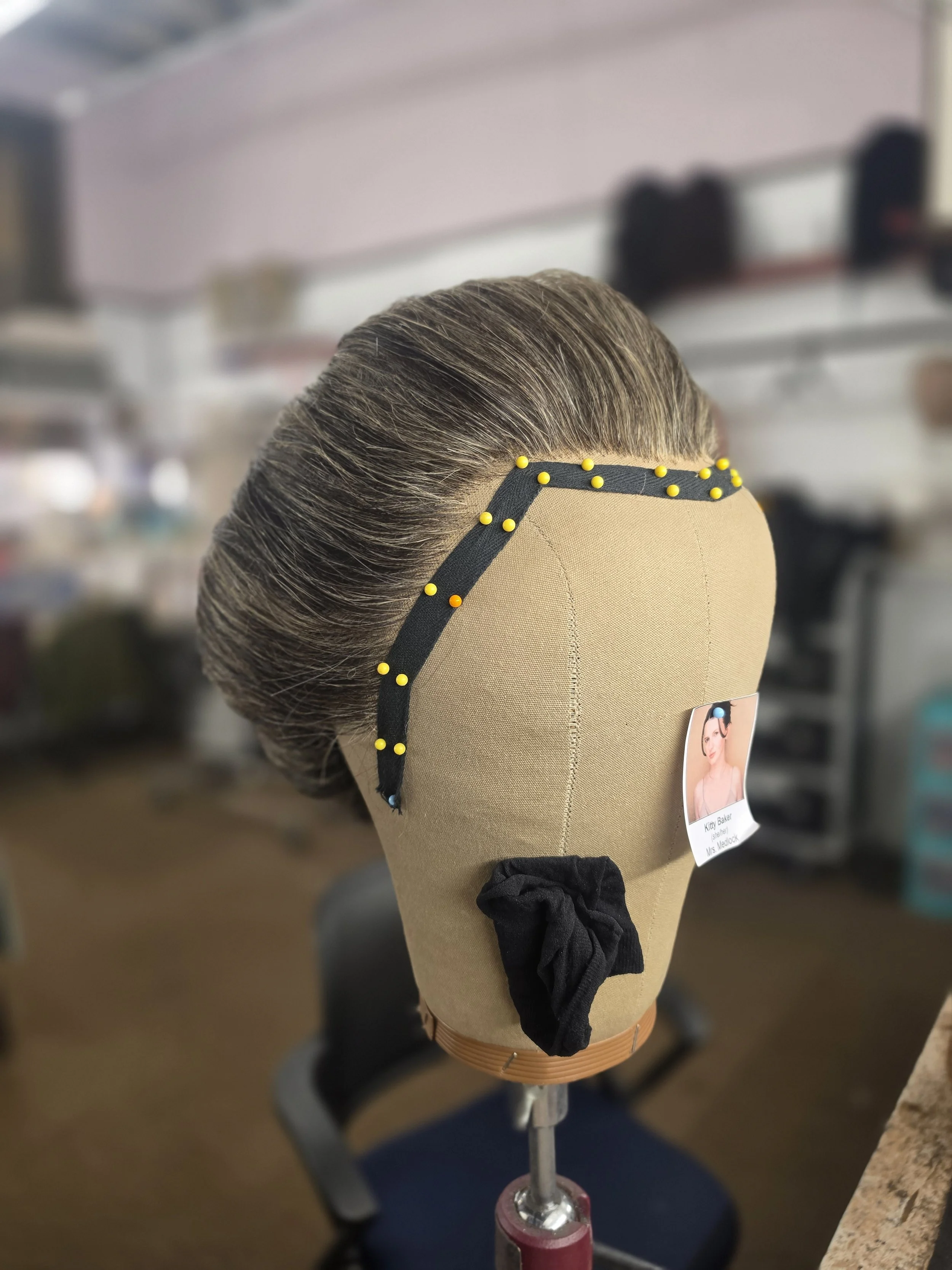 A mannequin head with gray hair, secured on a stand, with a photo ID tag that reads 'Kitty Baker, Streeter, Mrs. McClure.' The background shows a blurred workspace or salon environment.