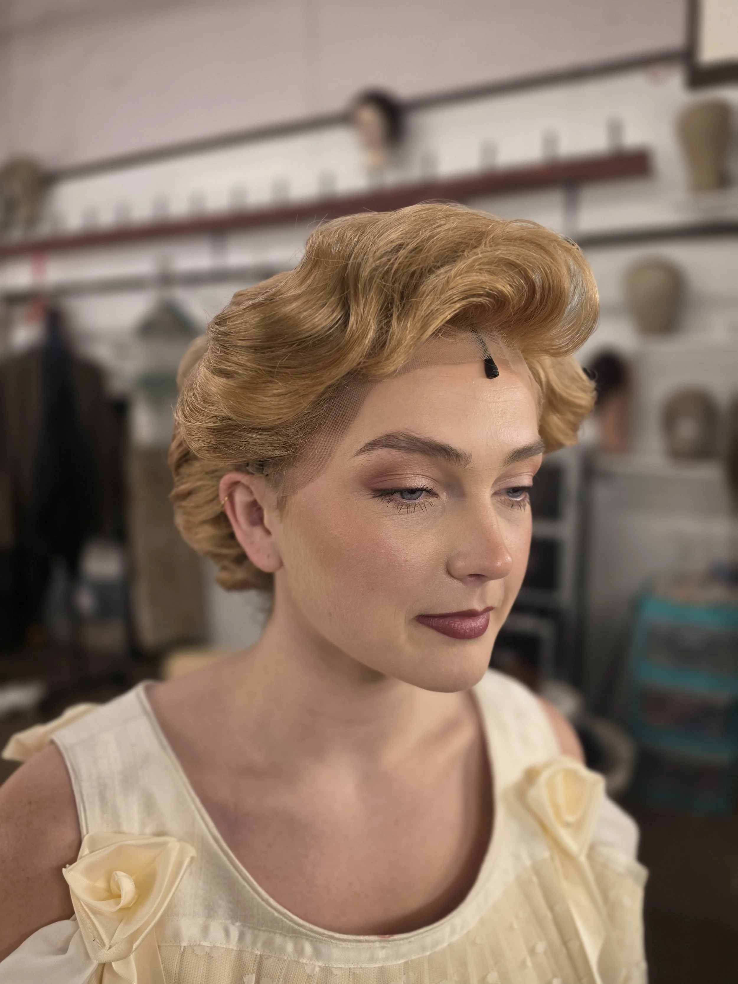 Close-up of a woman with vintage styled blonde hair, makeup, and a cream-colored dress with bow details on the shoulders, in an indoor setting.