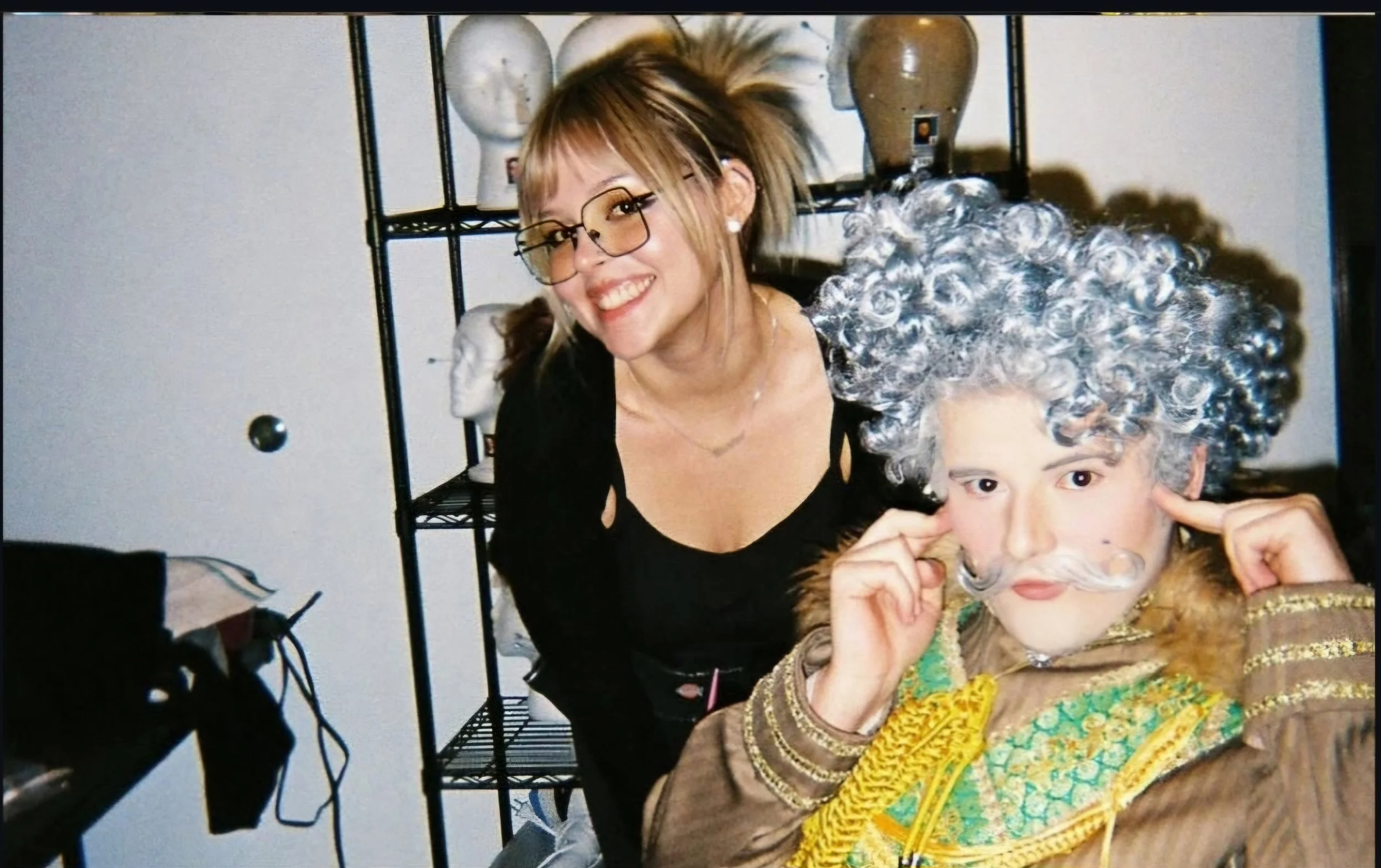 A woman with blonde hair and glasses laughing with a person dressed in a costume with a curly gray wig and makeup, possibly for a cosplay or costume event.