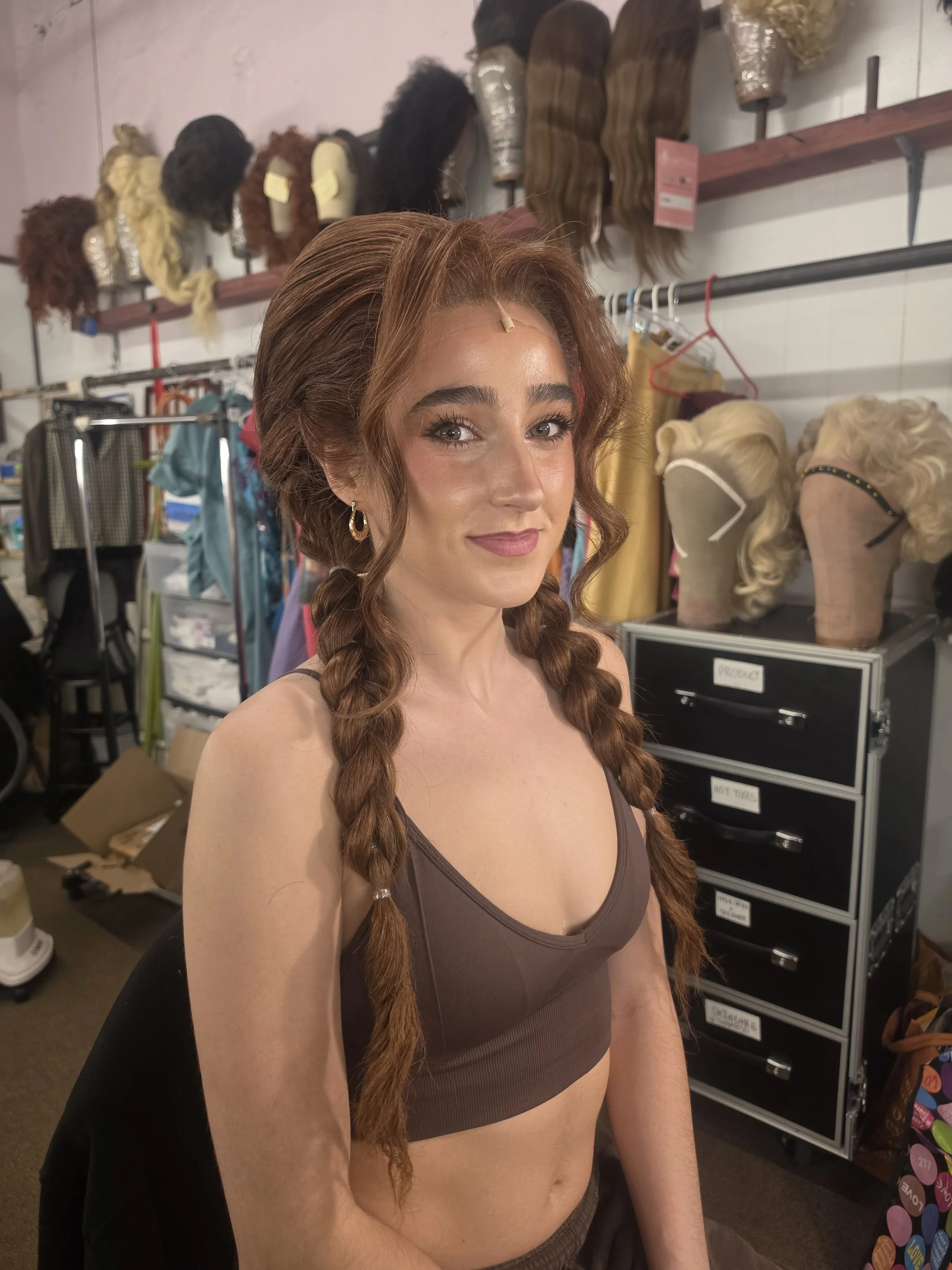 A woman with long, braided brown hair, wearing a brown sports bra, standing in a wig store with various wigs and wig mannequins in the background.