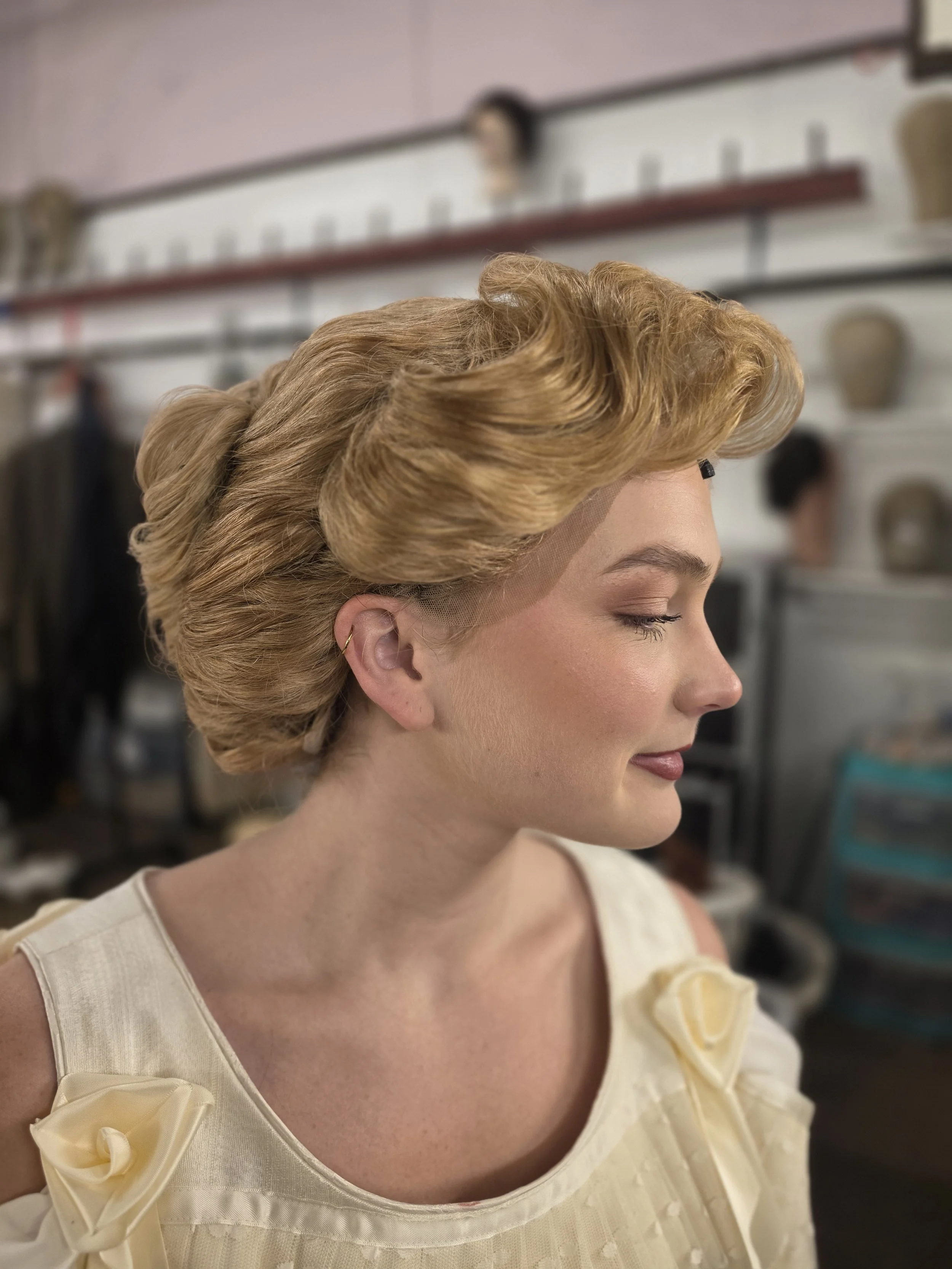 Side profile of a woman with vintage blonde hair styled in large waves, wearing a cream-colored dress with ribbon detail on the shoulder.