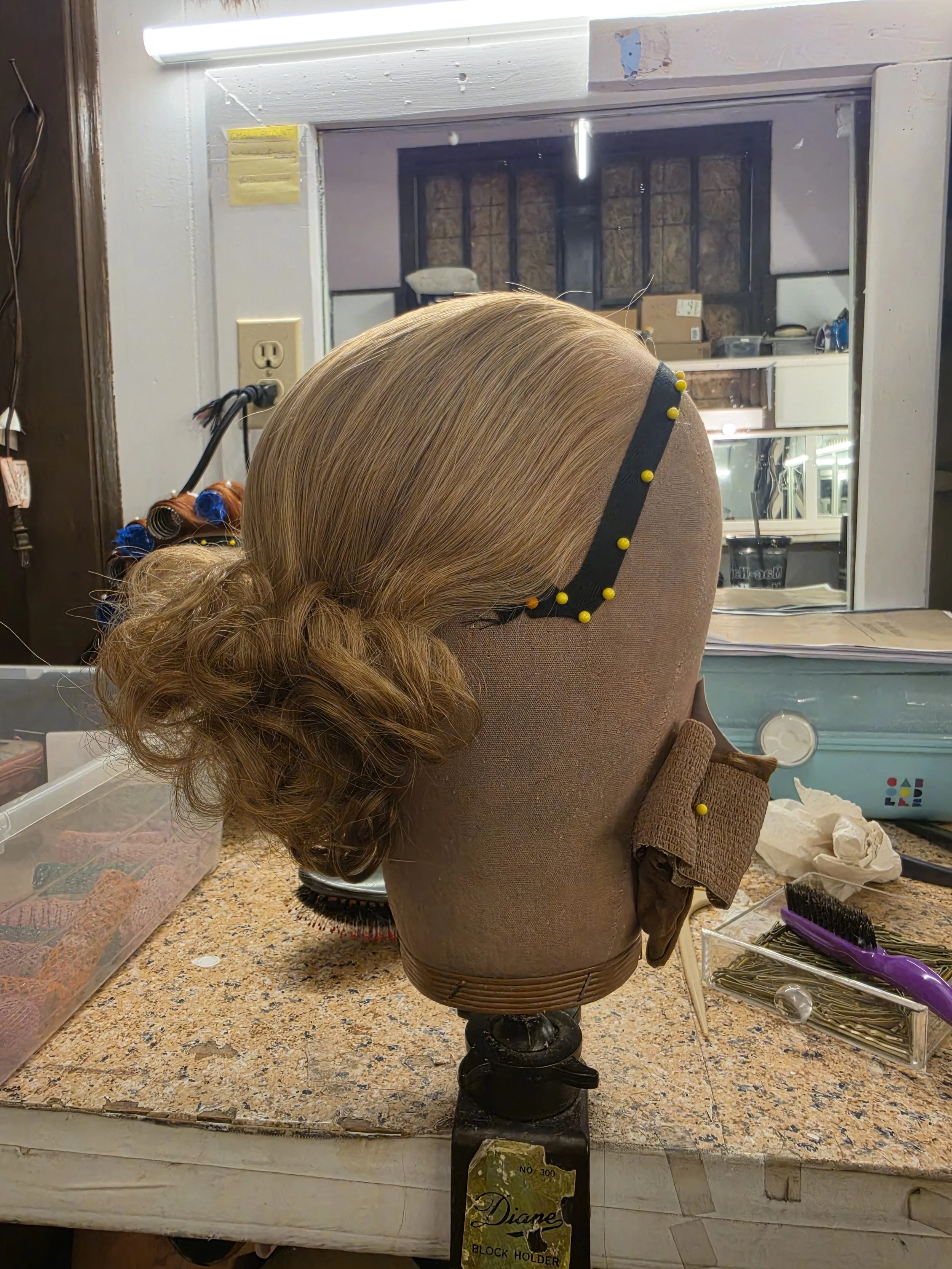 A mannequin head with styled, curly, red hair pinned with blue curlers, placed on a workbench in a salon or makeup studio with various tools and supplies around.