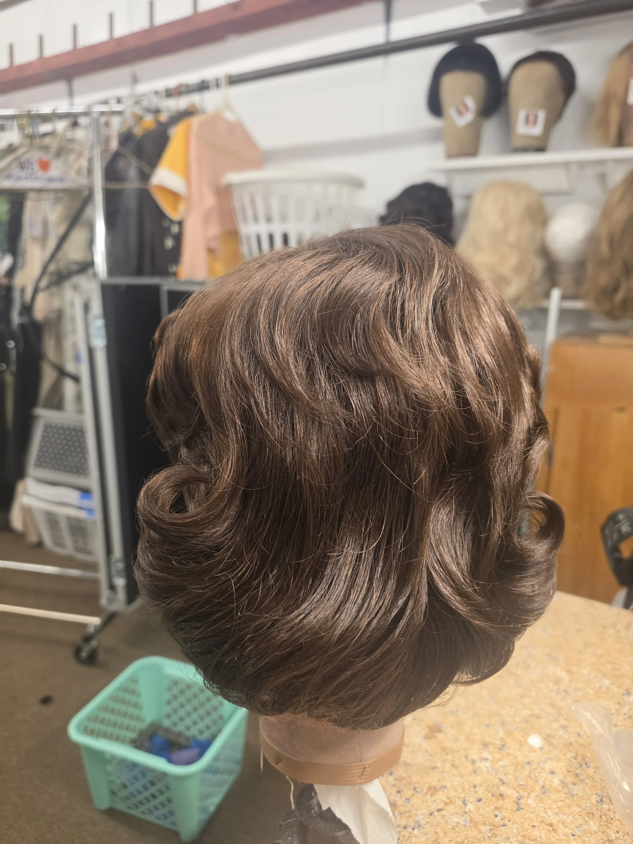 A mannequin head with styled brown hair, set on a stand in a store or salon, with hairdressing supplies and shelves in the background.