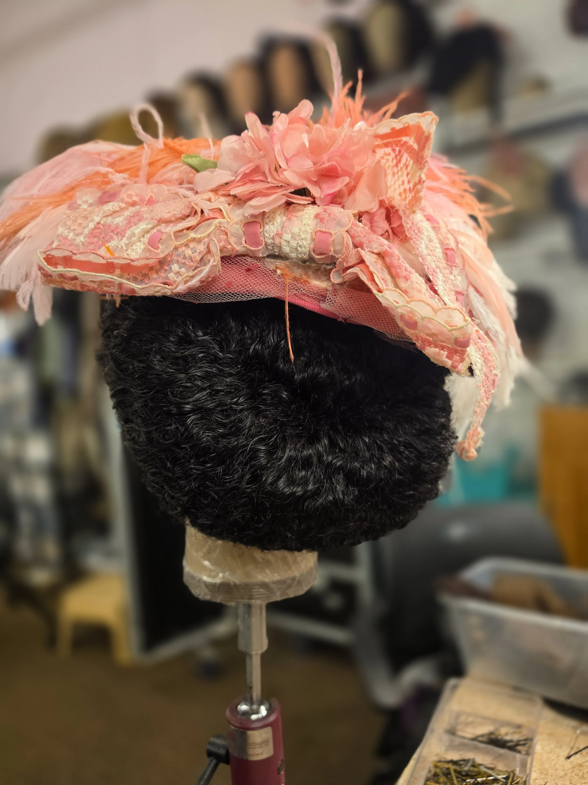 The image shows a mannequin head with black hair wearing a decorative pink hat made of fabric, flowers, and lace, positioned on a stand in a workspace.
