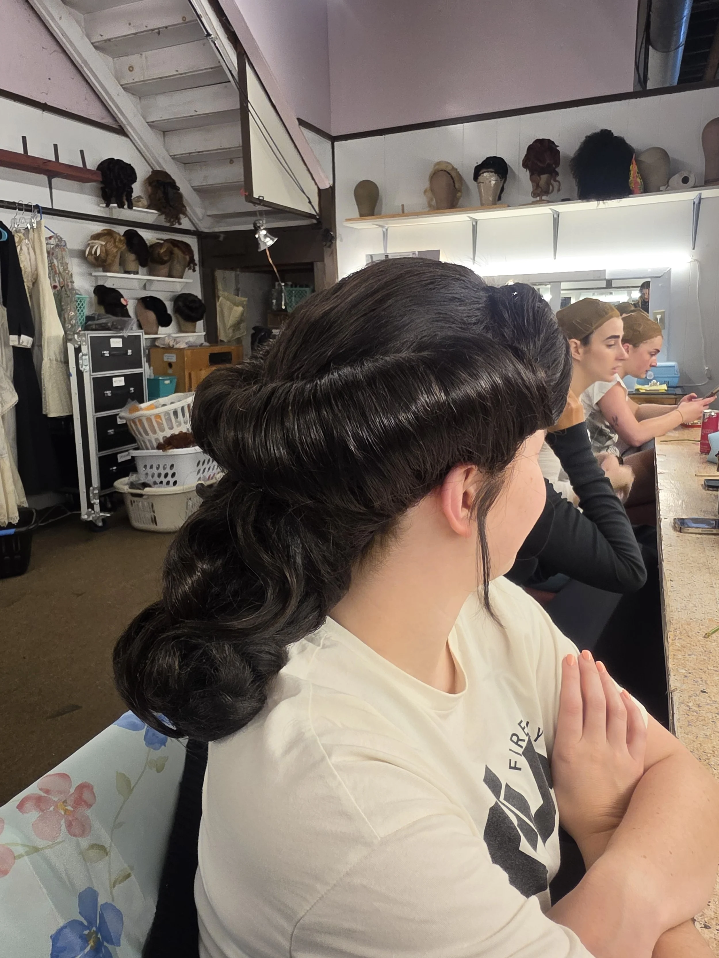 People sitting at a bar or table in a salon or beauty shop with wig displays on shelves in the background.