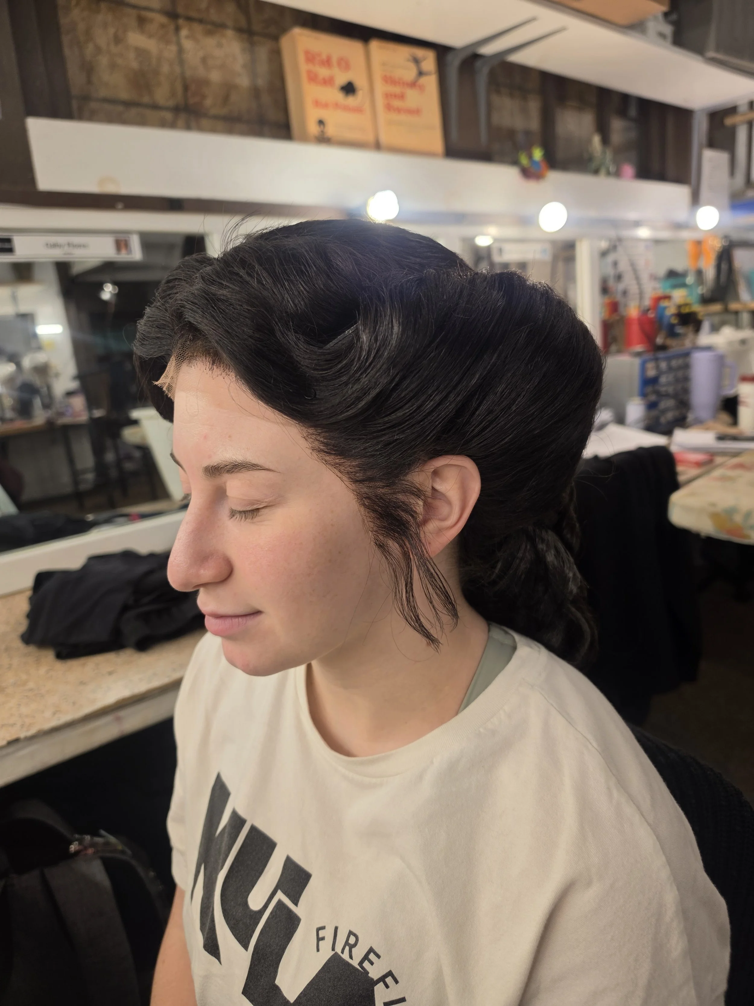 A woman with dark, vintage styled hair sitting in a salon or studio environment, with hair rollers visible and hair partially styled.