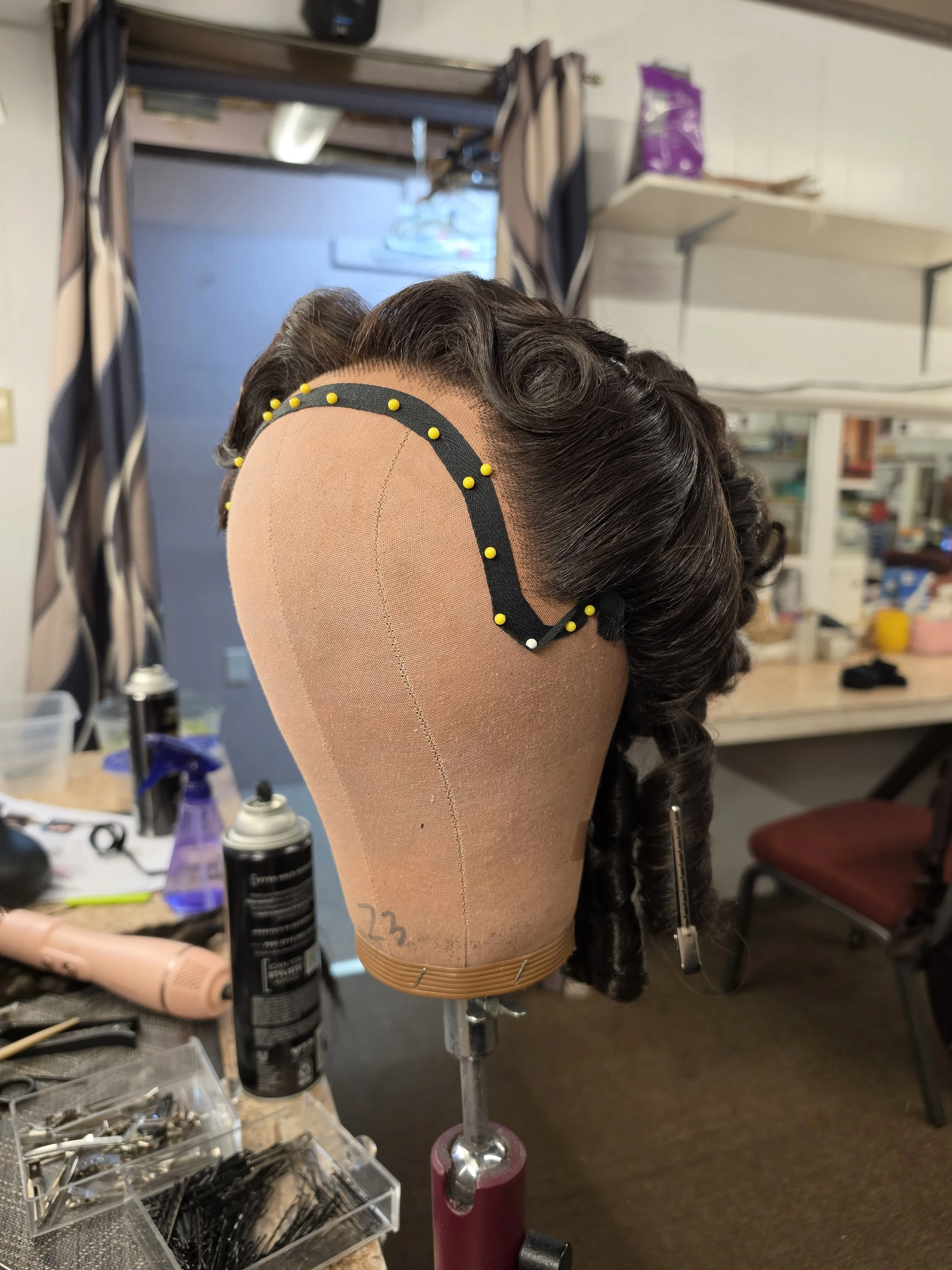 A mannequin head on a stand decorated with a vintage hairstyle wig, with a black headband secured with yellow pins. The background shows a cluttered workspace with sprays, tools, and fabric, suggesting a wig or hairstyling station.