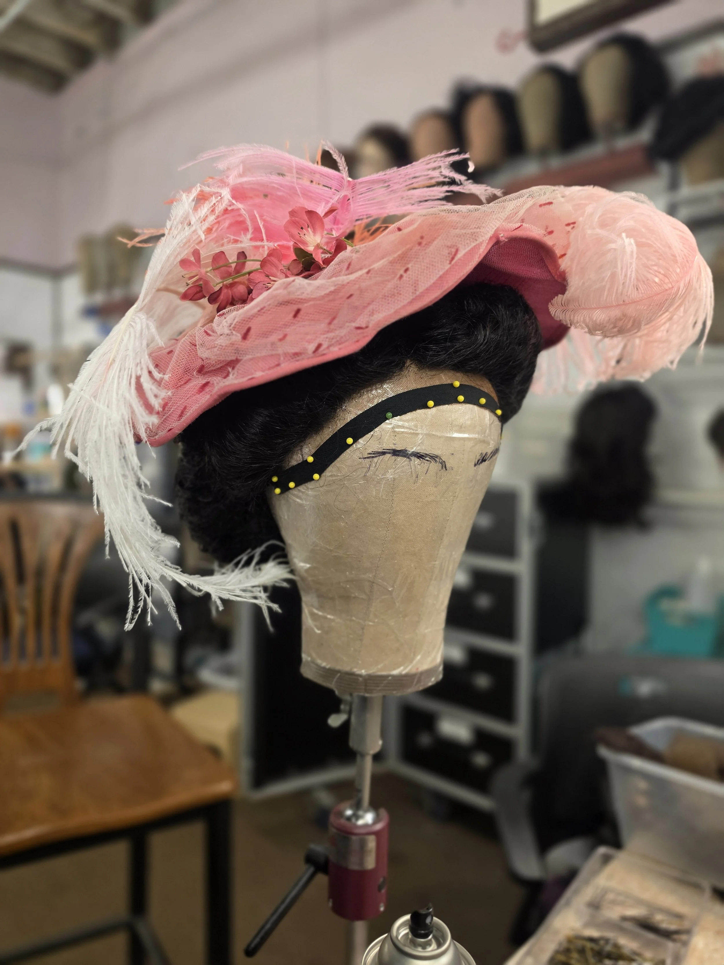 A mannequin head with black hair wearing a pink hat decorated with pink flowers and feathers, mounted on a stand in a workshop.