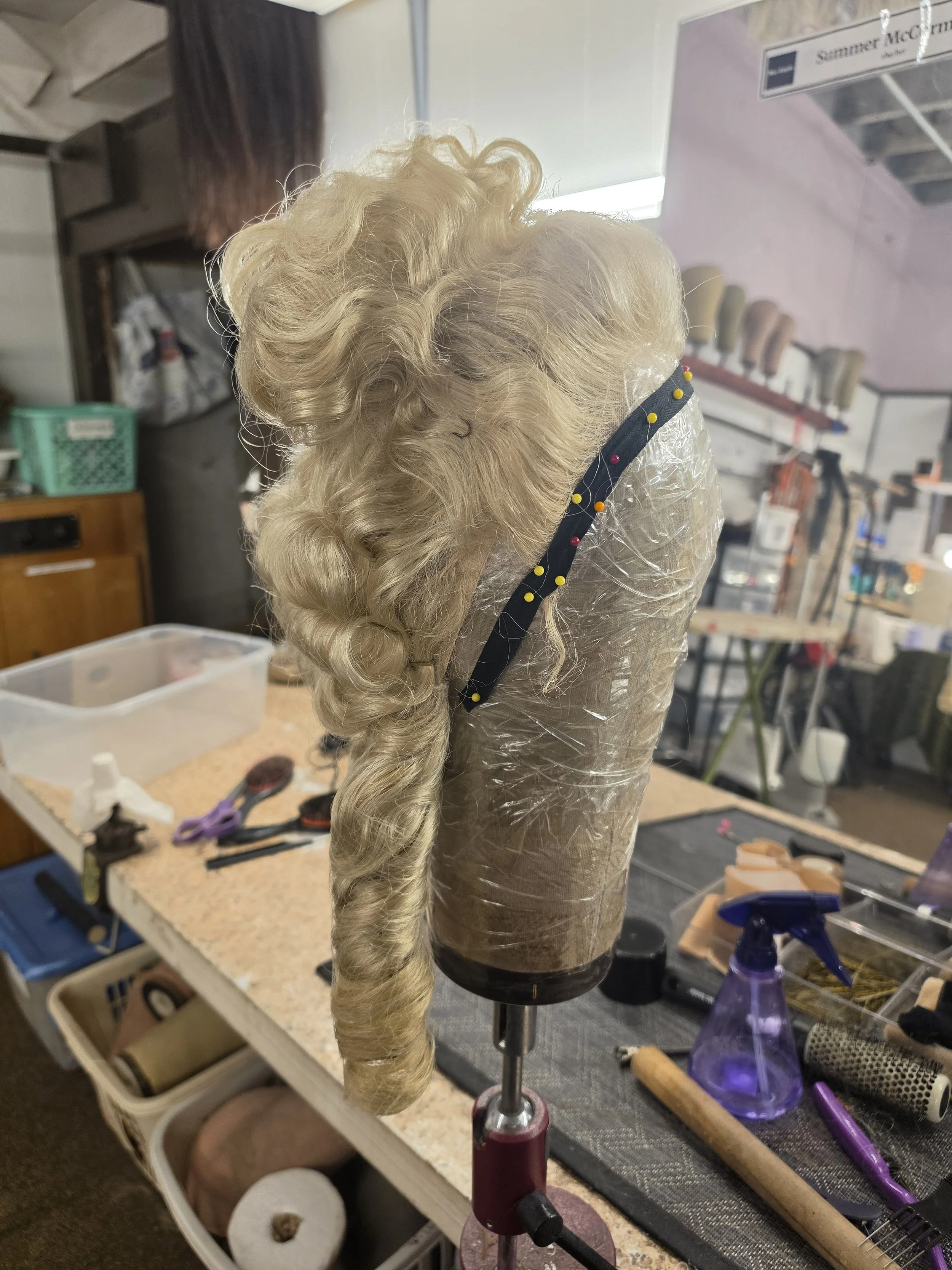 A blonde, curly wig on a mannequin head atop a worktable with hairstyling tools and supplies, including scissors, brushes, a spray bottle, and rolls of material in the background.