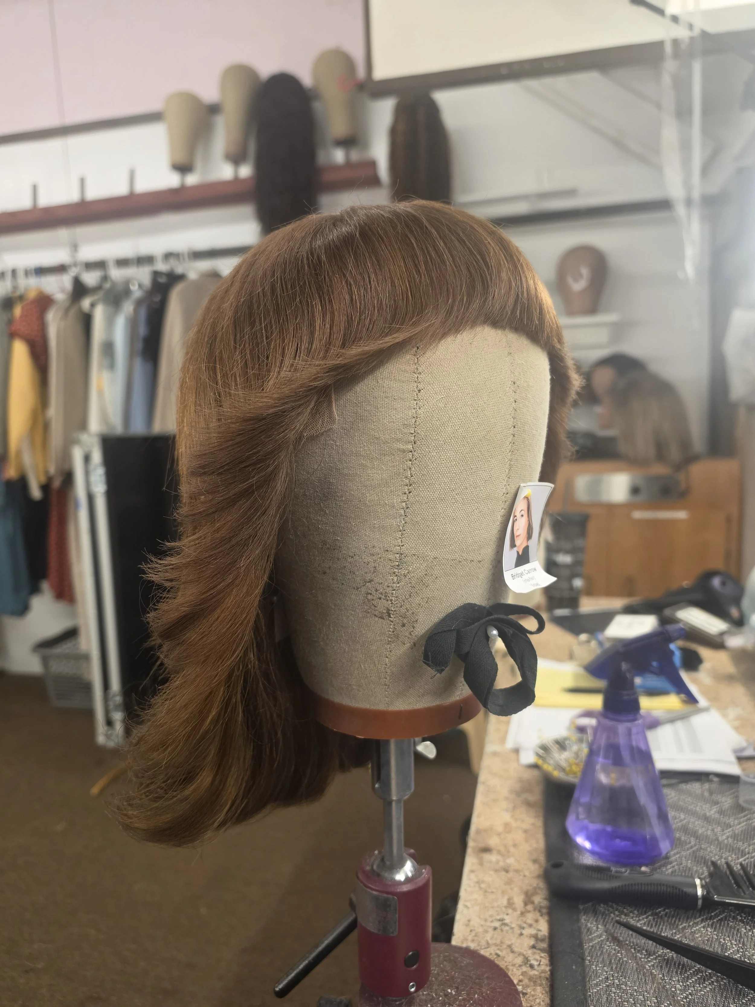 A mannequin head with a long, wavy, auburn wig placed on a work table in a hair salon or wig studio. The background shows shelves with hairpieces and salon tools.