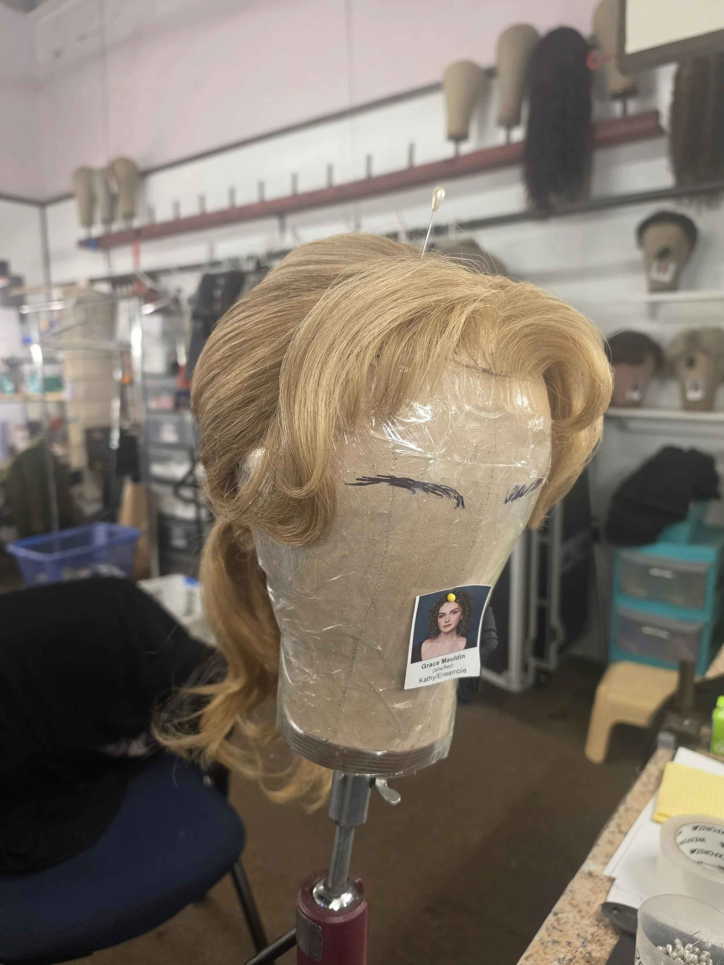 A blonde wig with curls on display on a mannequin head, secured on a stand in a wig shop.