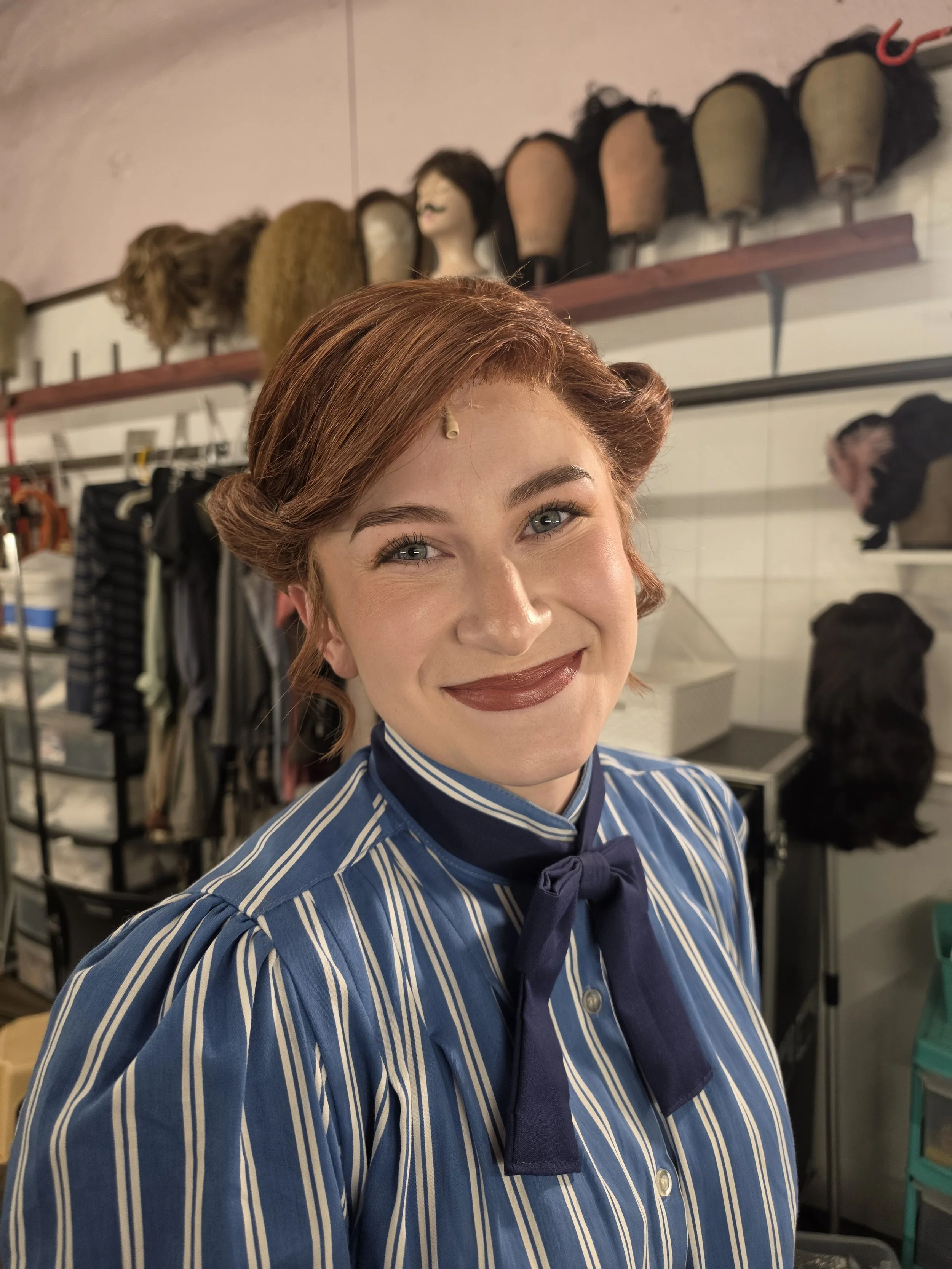 A woman with red hair styled in vintage waves, wearing a blue and white striped blouse and a navy bow tie, smiling in a hair salon with wig heads on shelves behind her.