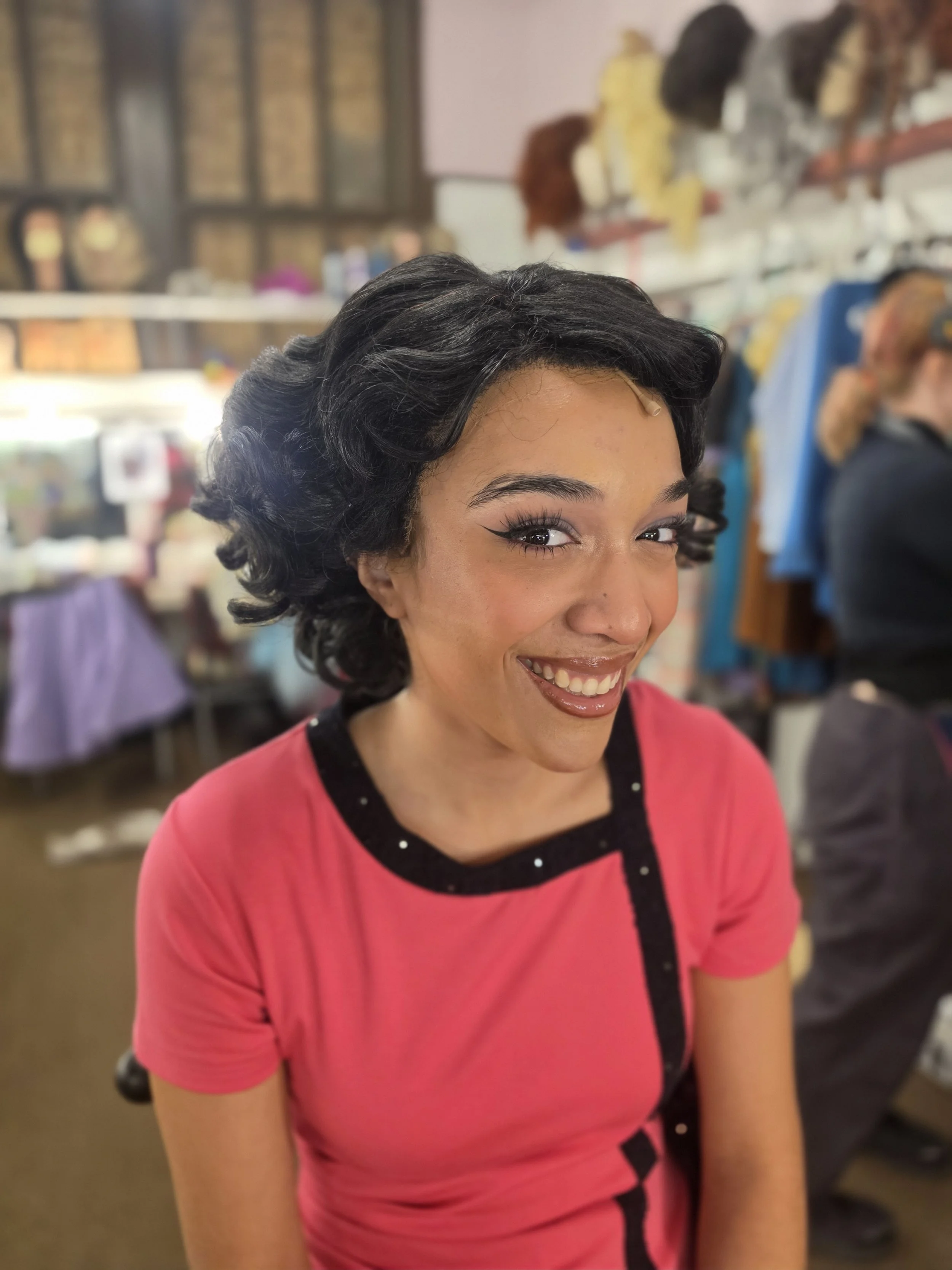 A woman with dark, curly hair and a bright pink shirt, smiling, in a room with shelves and various objects in the background.