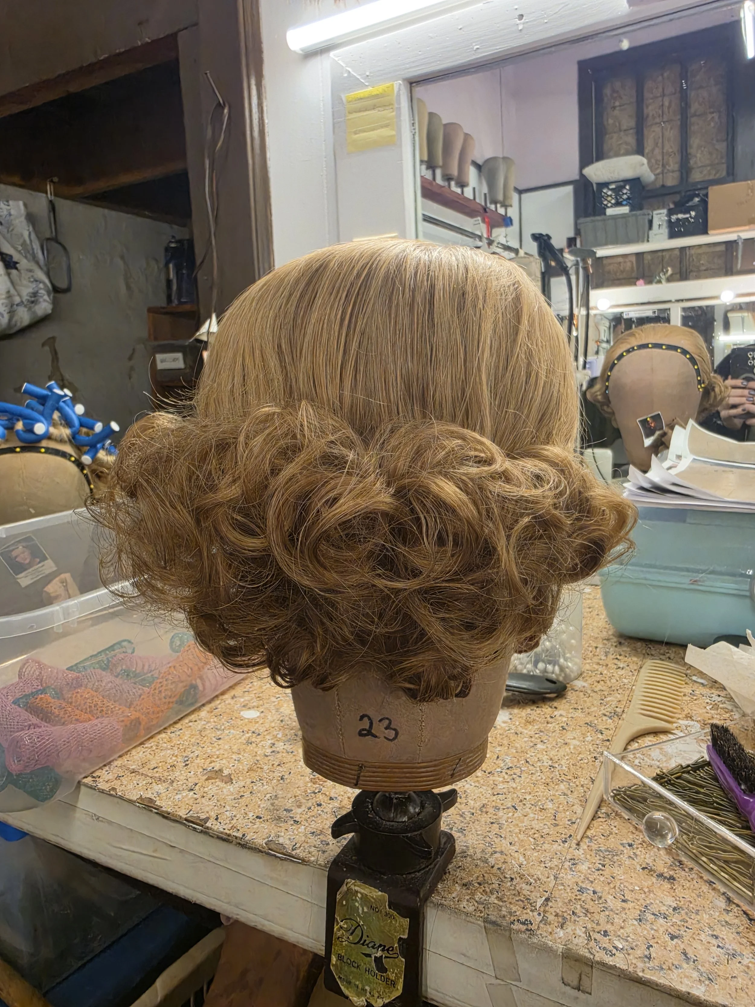 A mannequin head with curly, red hair on a wooden stand. Behind it, a mirror reflects the room with various hairdressing tools and supplies, including hair curlers, brushes, and clips, on a cluttered table and shelves.