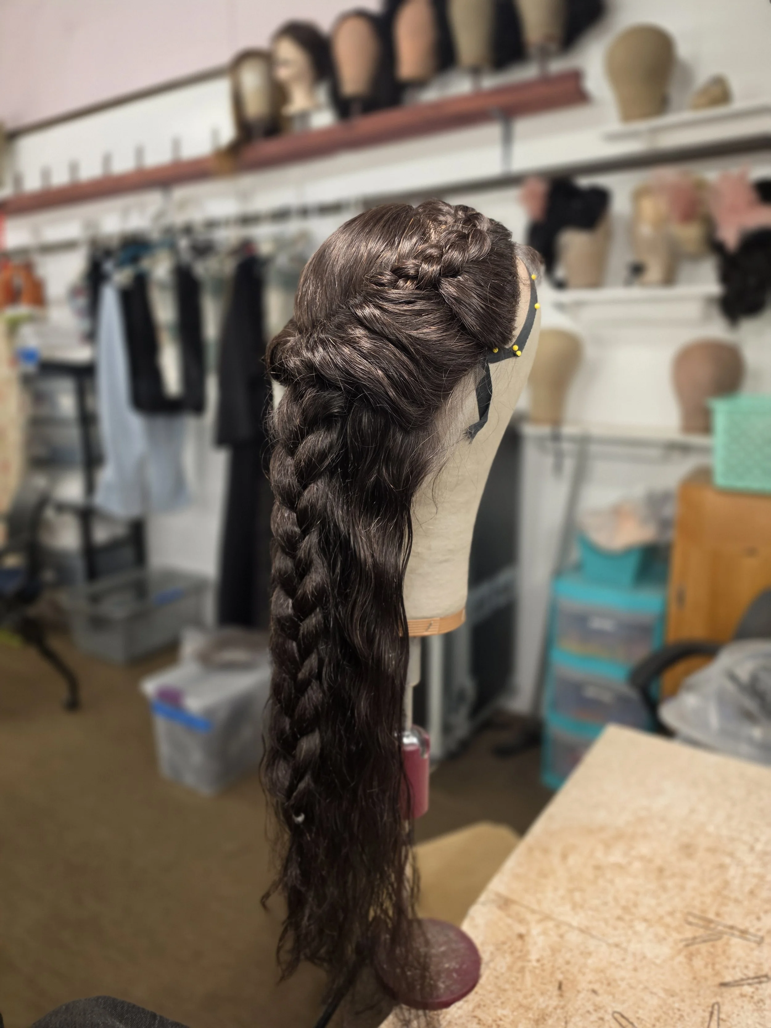 A mannequin head on a stand with a long dark brown braid, surrounded by hairdressing tools and hairpieces in a salon or costume shop.