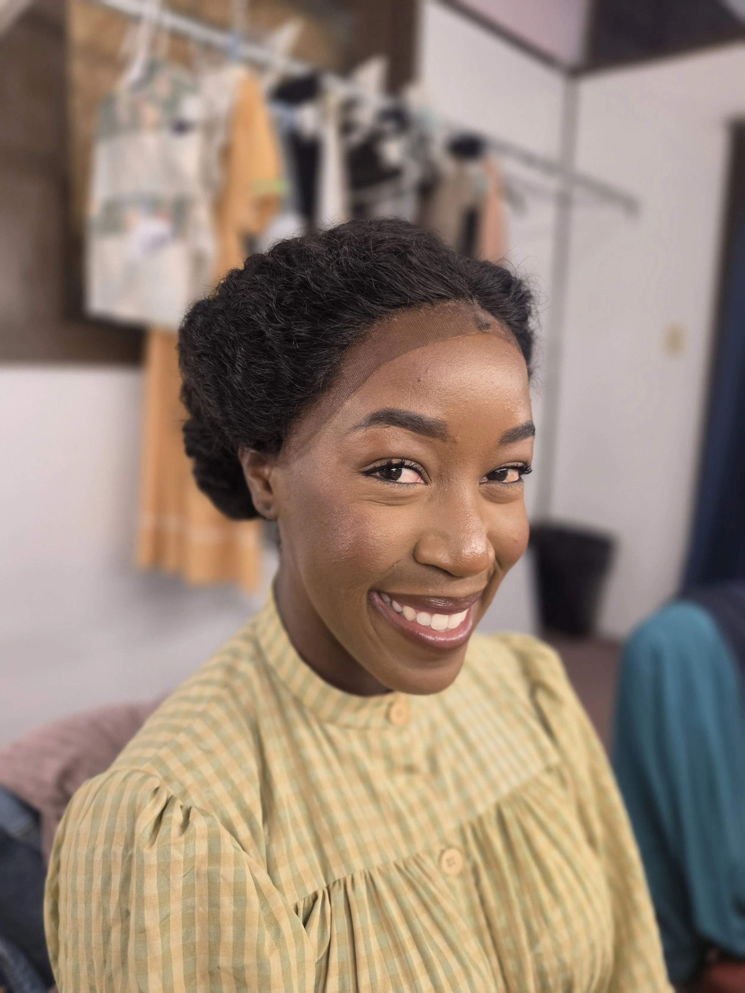 A smiling woman looking at the camera, wearing a yellow striped shirt, with a blurred background of clothing hanging on a rack.