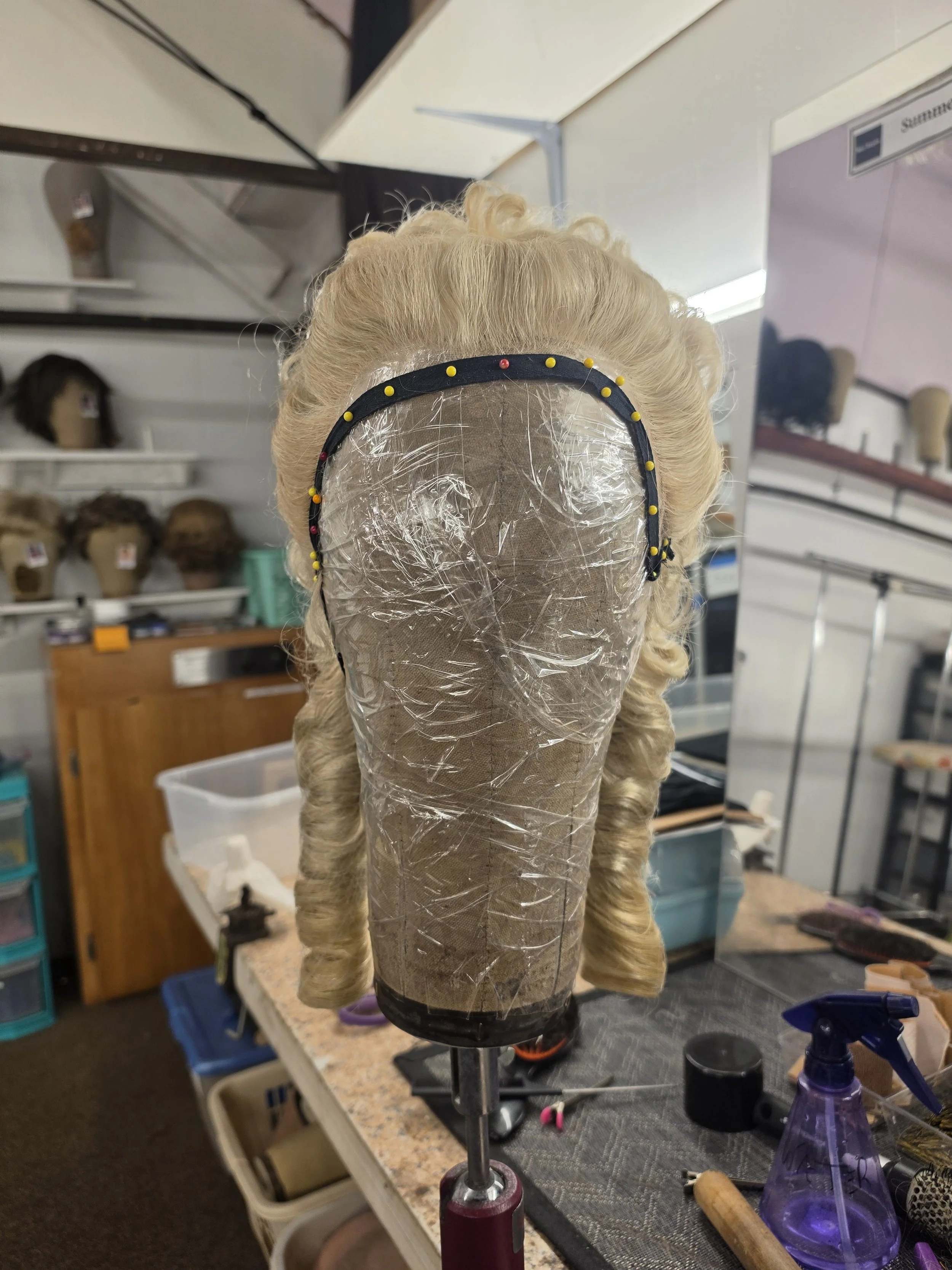 A wig stand with a blonde, curly wig in a wig making or styling workshop. The background shows shelves with mannequin heads and wig styles, workshops tools, and supplies on a workbench.