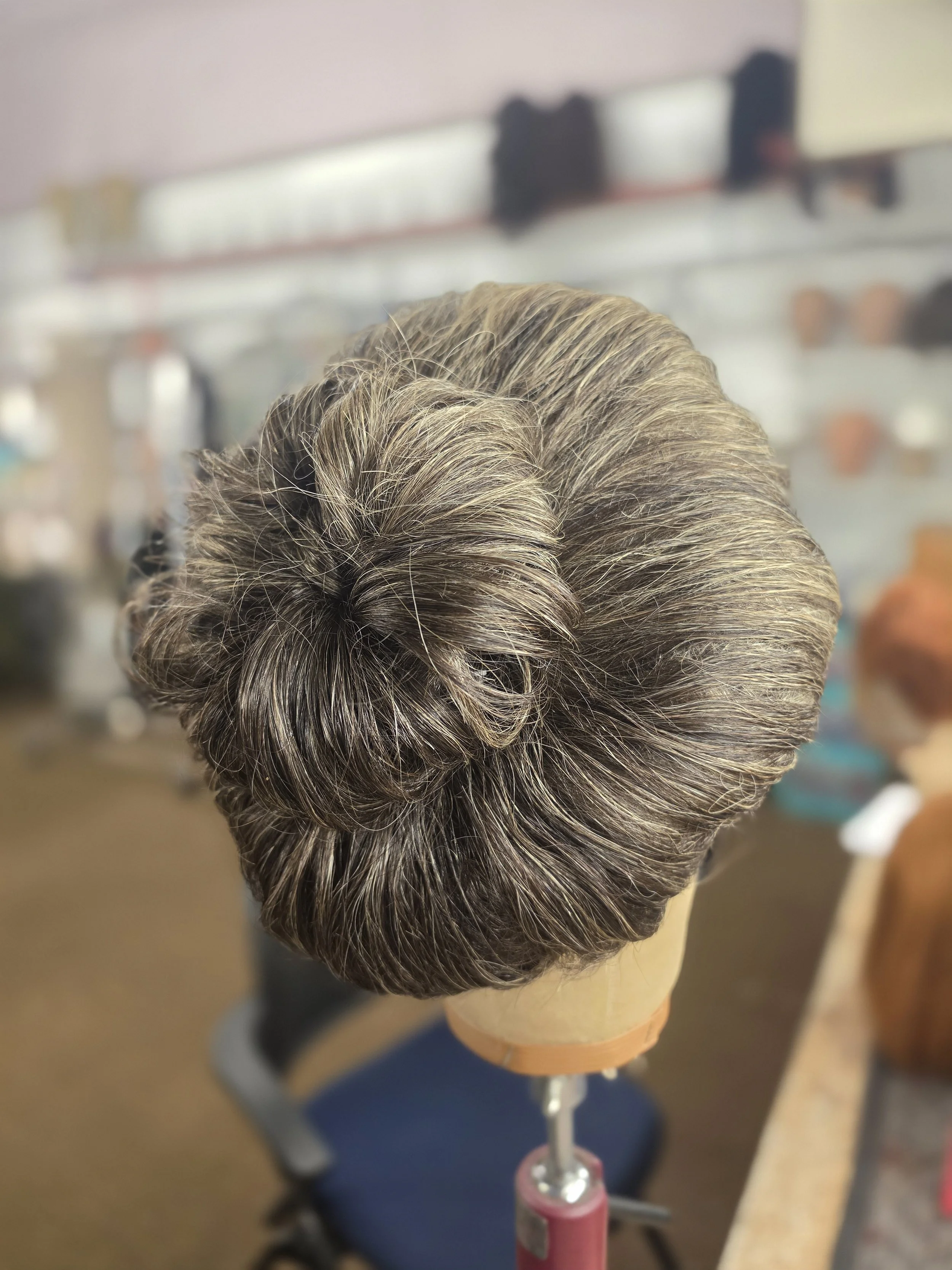 A mannequin head with styled, gray and white hair, mounted on a pink stand in a salon or barber shop, with shelves of hair products in the background.