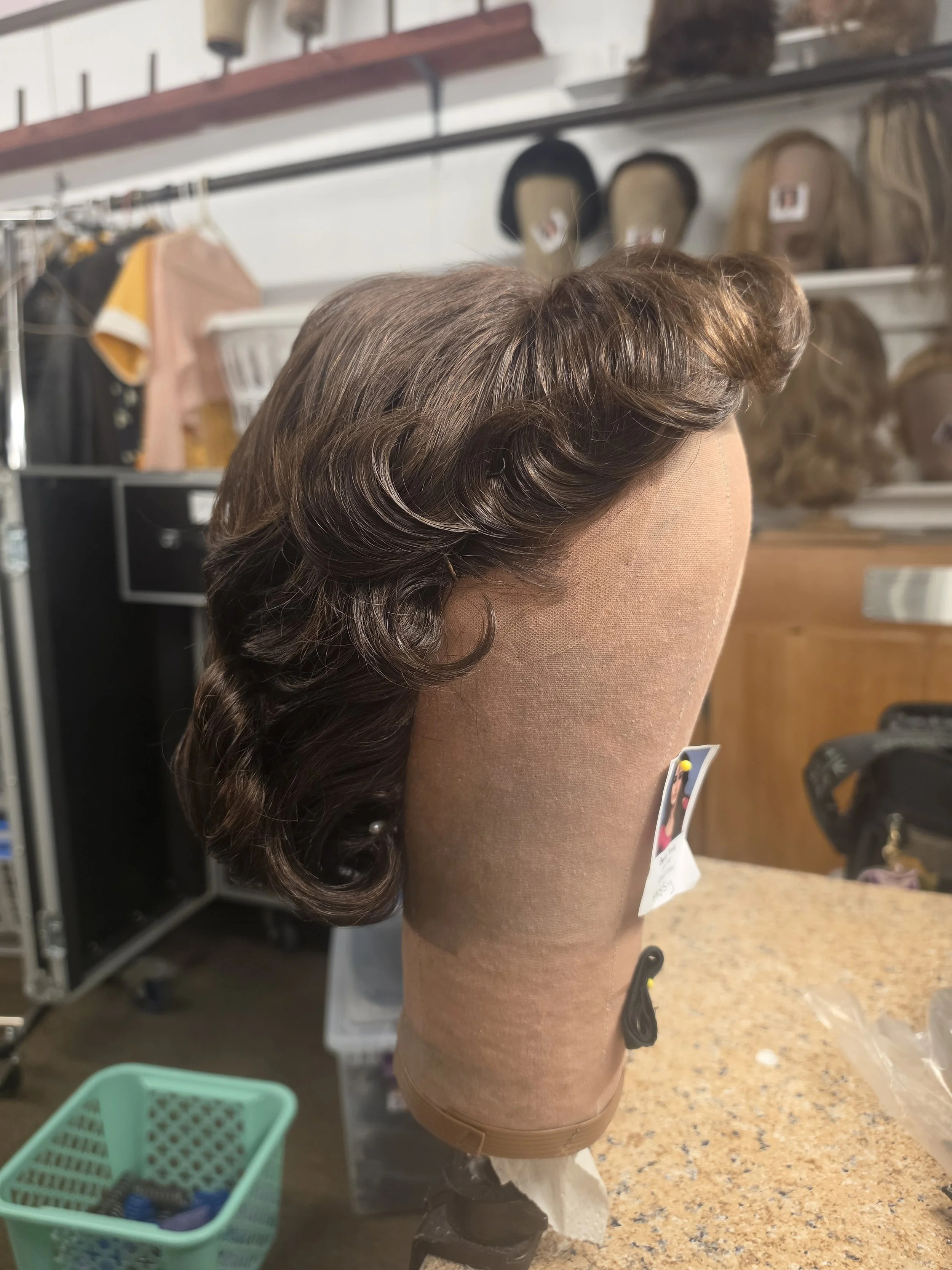 A mannequin head with styled, wavy brown hair, positioned on a countertop in a room with shelves holding bags and shoes in the background.