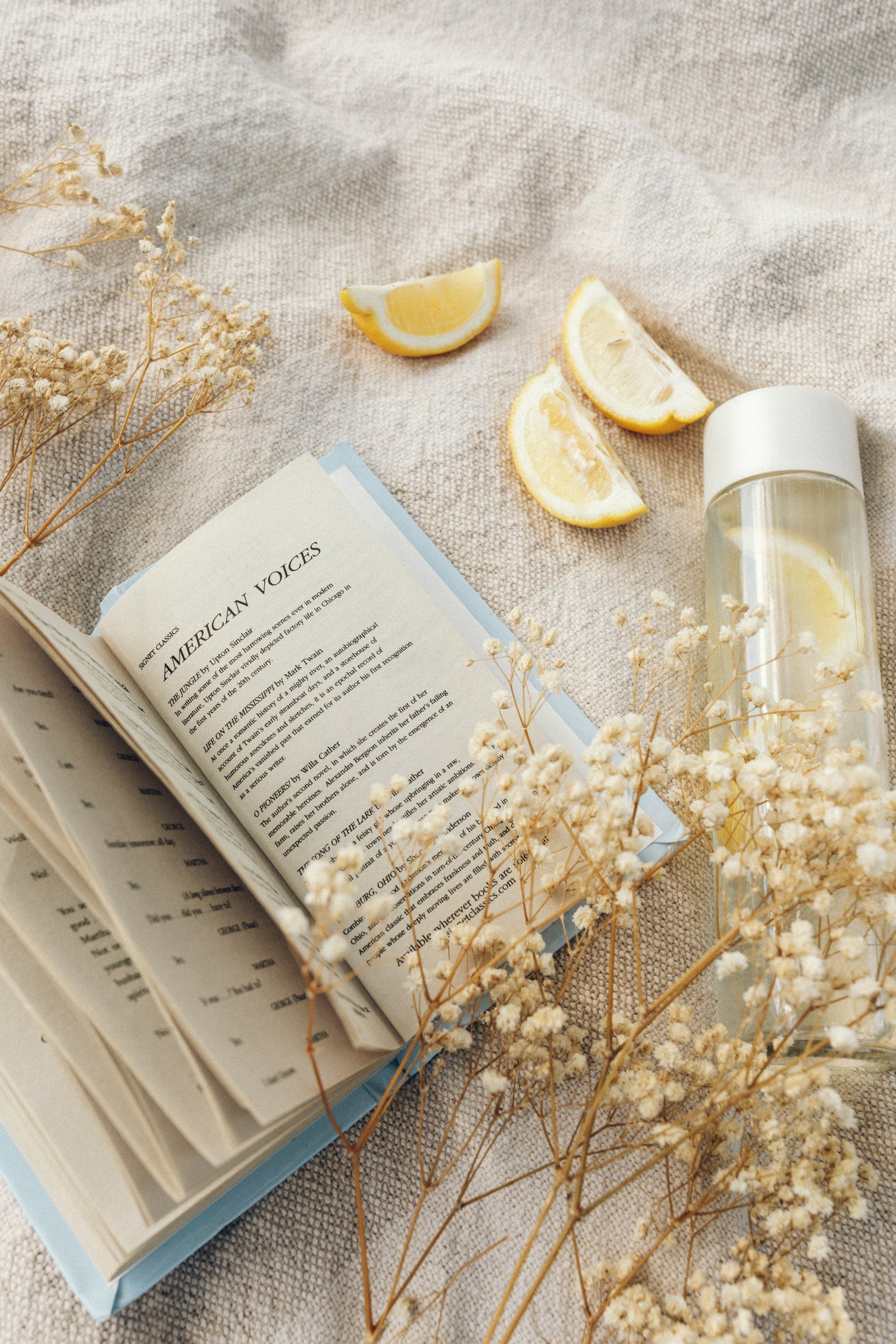A book open on a beige textured fabric surface with dried white flowers and lemon wedges, and a glass container with water and lemon slices.